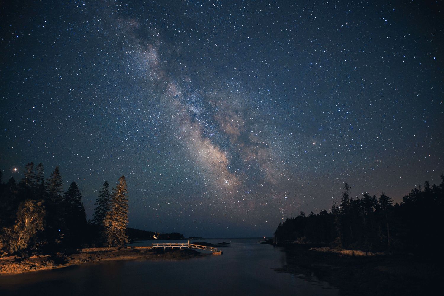Night sky over a dark, calm body of water, with trees on either side. The Milky Way galaxy is visible.