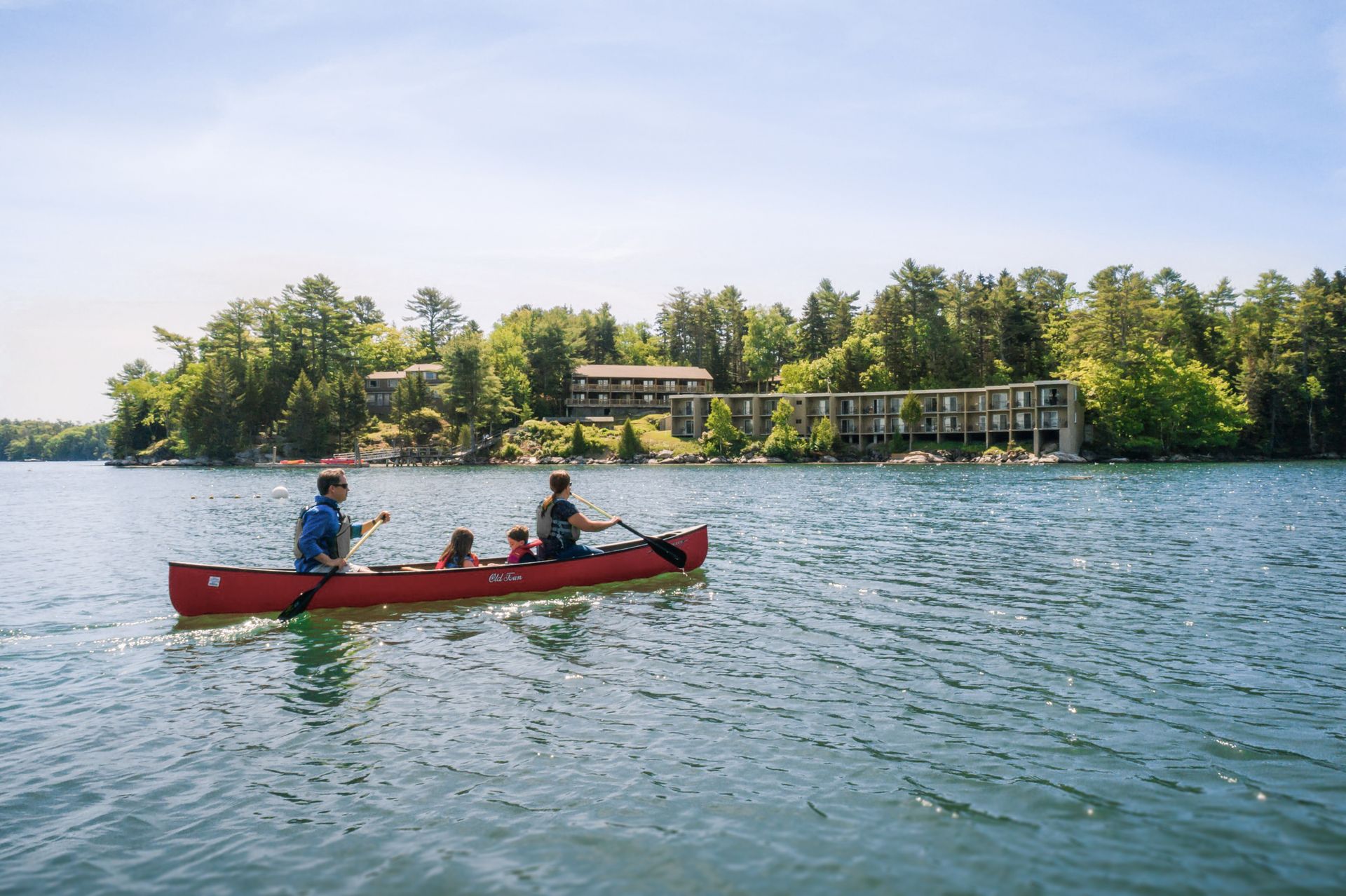 People canoeing on a lake, buildings and trees in the background.