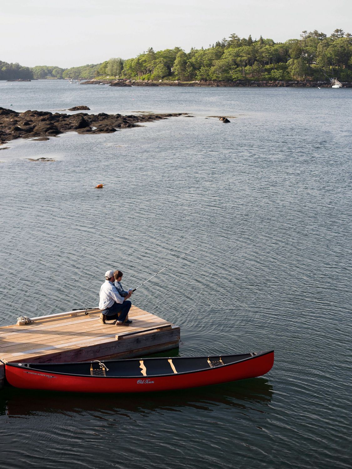 Two people fish off a dock near a red canoe on a lake with a wooded shoreline.