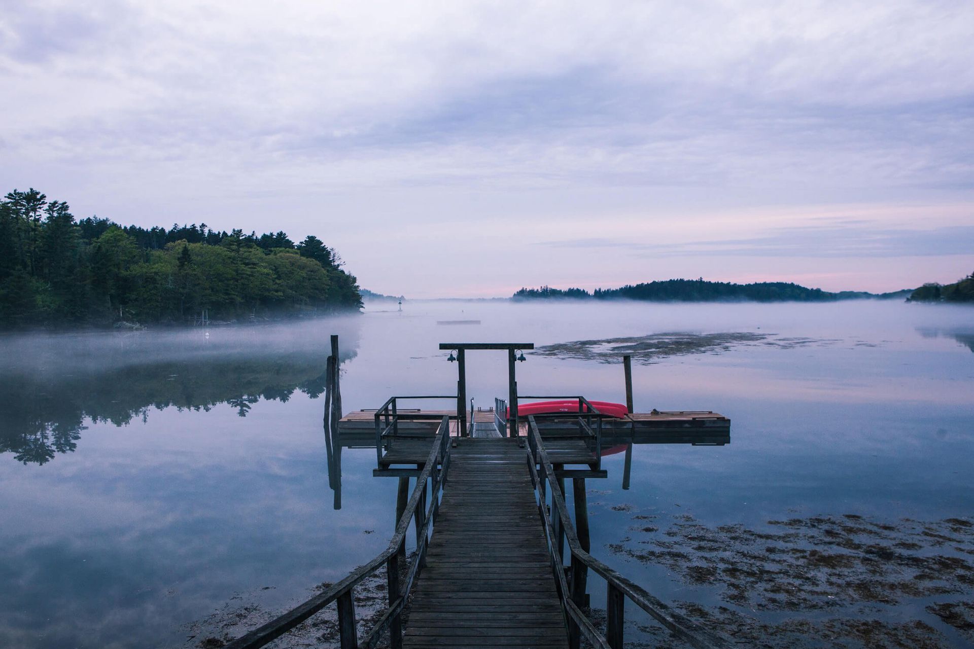 Wooden dock extending over calm water at dawn. Mist, trees, and cloudy sky reflected in the still lake.