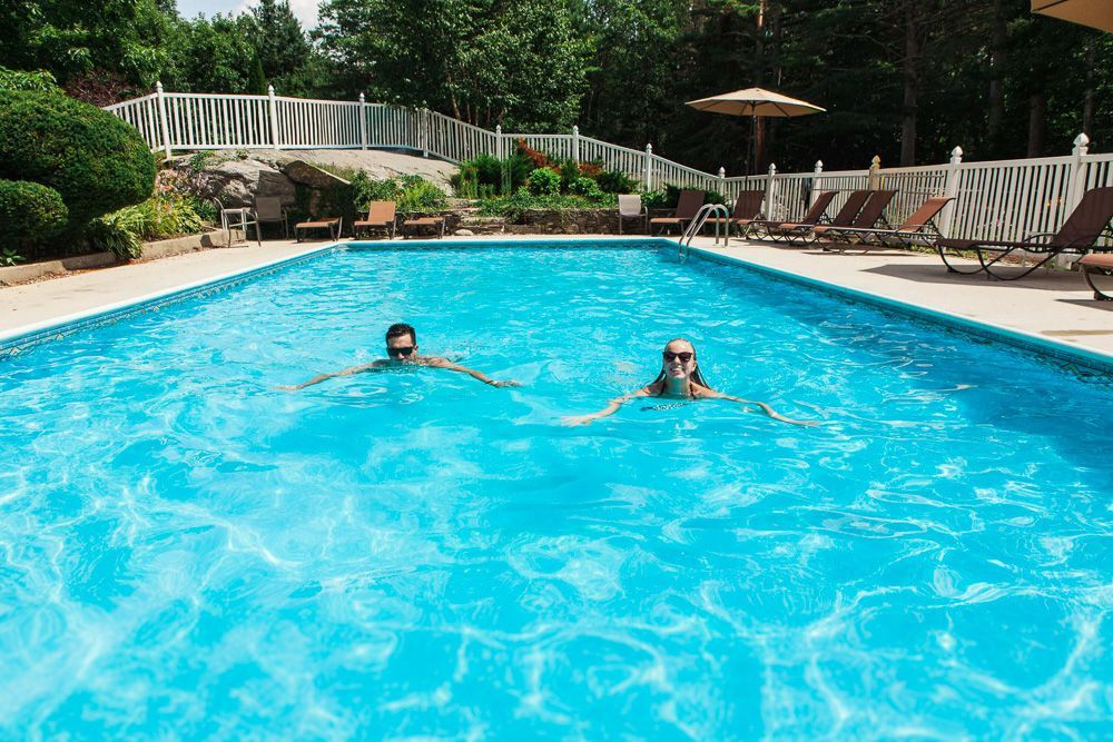 Two people swimming in a blue pool on a sunny day near lounge chairs and white fence.