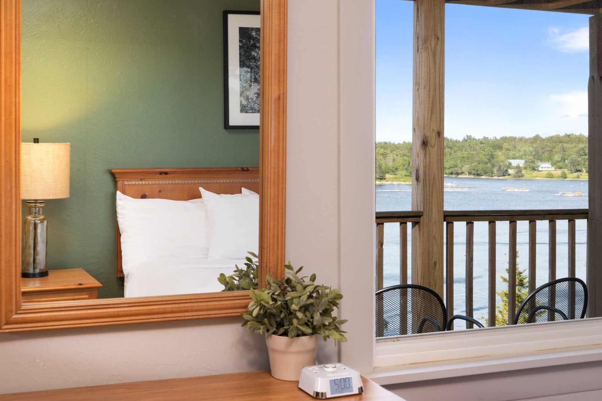 Bedroom view: Mirror reflects bed, bedside table. Window shows lake and balcony with chairs.