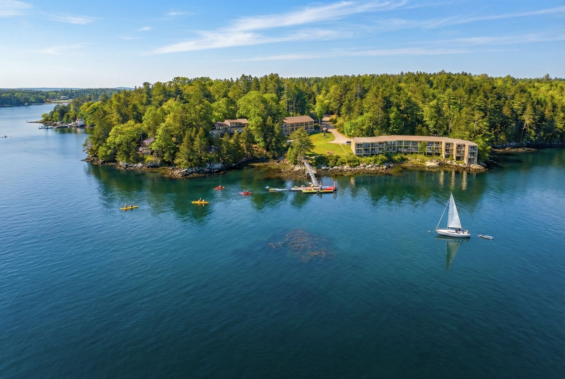 Aerial view of island with buildings, surrounded by blue water; boats and kayaks visible.