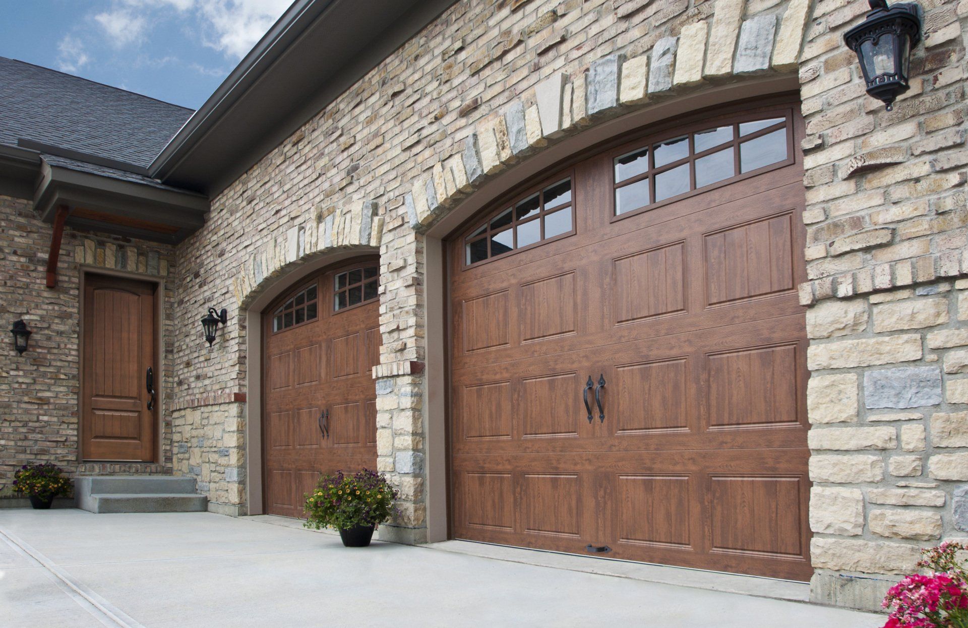 Two brown arched garage doors and a wooden door with stone facade.