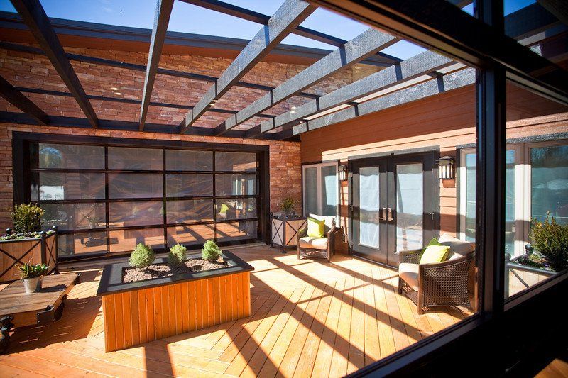 Outdoor patio with wooden deck, brick wall, and glass garage-style door and roof.  Chairs and a planter are visible.