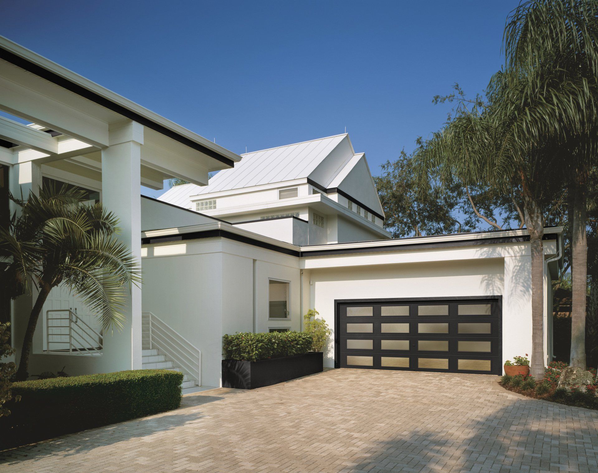 Modern white house with a black-framed glass garage door. Gravel driveway, green shrubbery, and a blue sky.
