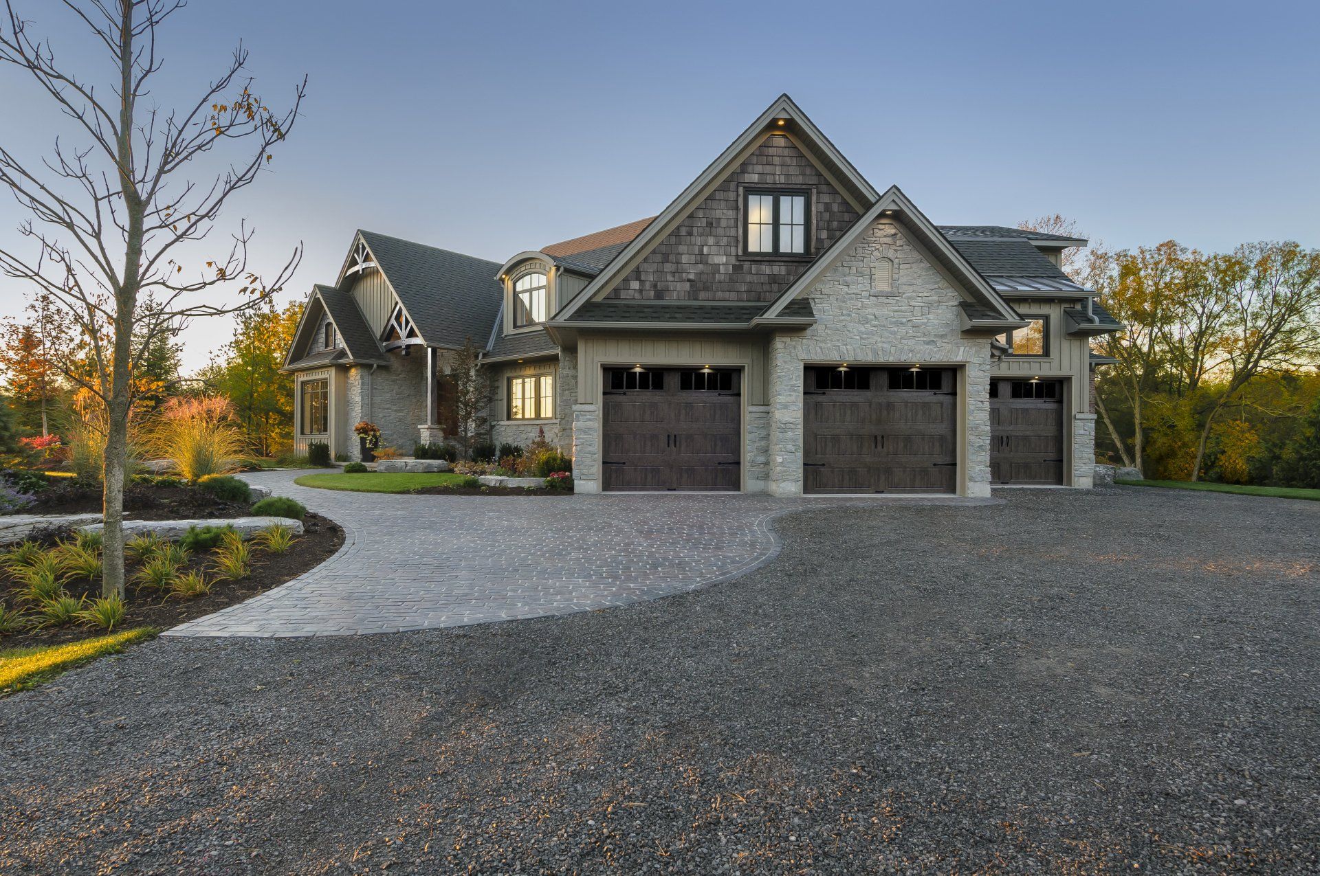 Gravel driveway leading to a two-story house with stone and wood siding, three garage doors, and a clear blue sky.