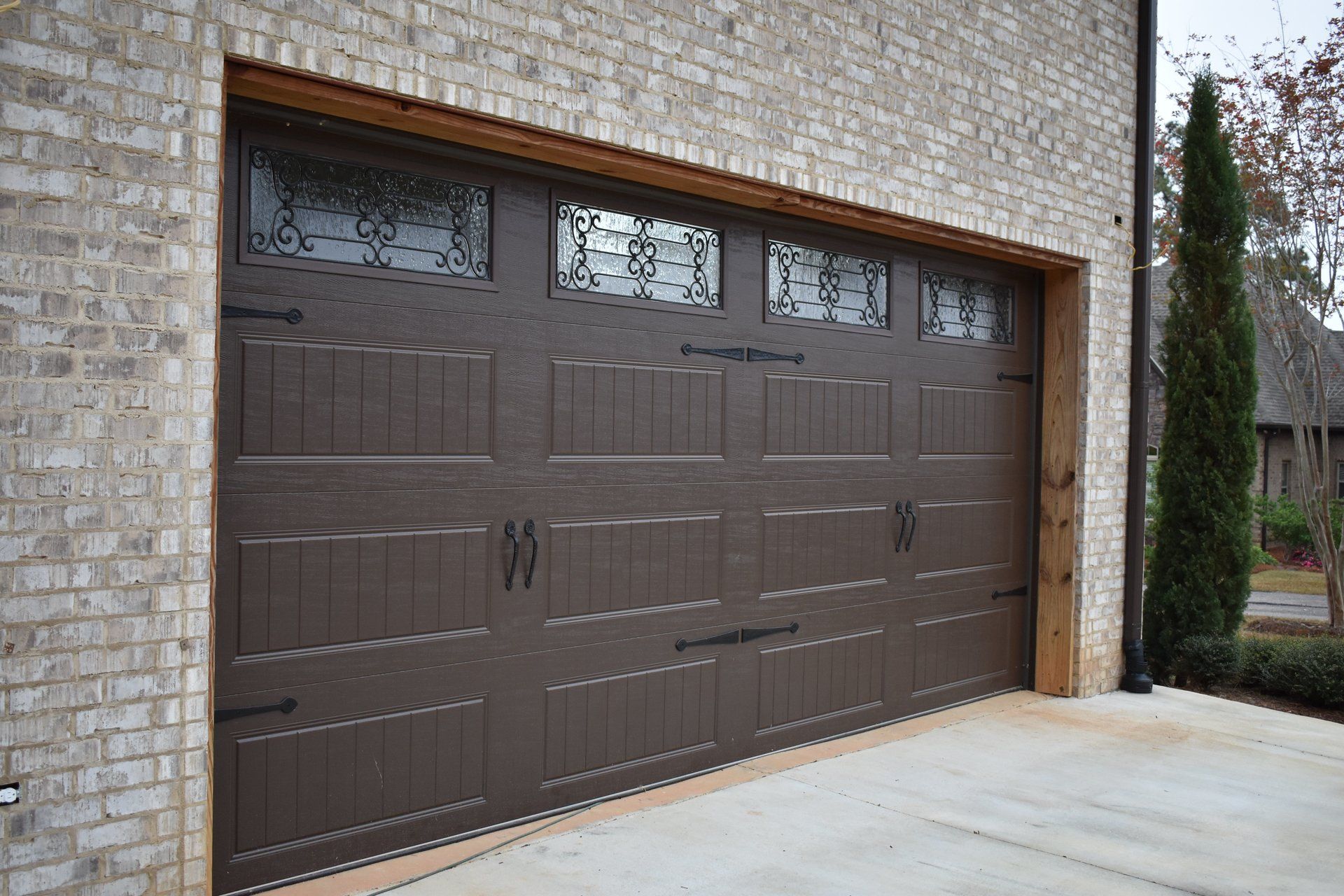 Brown garage door with decorative glass panels, hardware, and textured panels. Set in a brick building.
