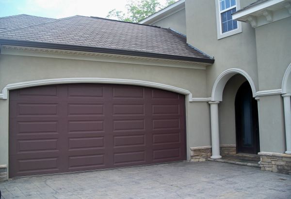 Brown garage door on a beige house with a curved top. Concrete driveway.