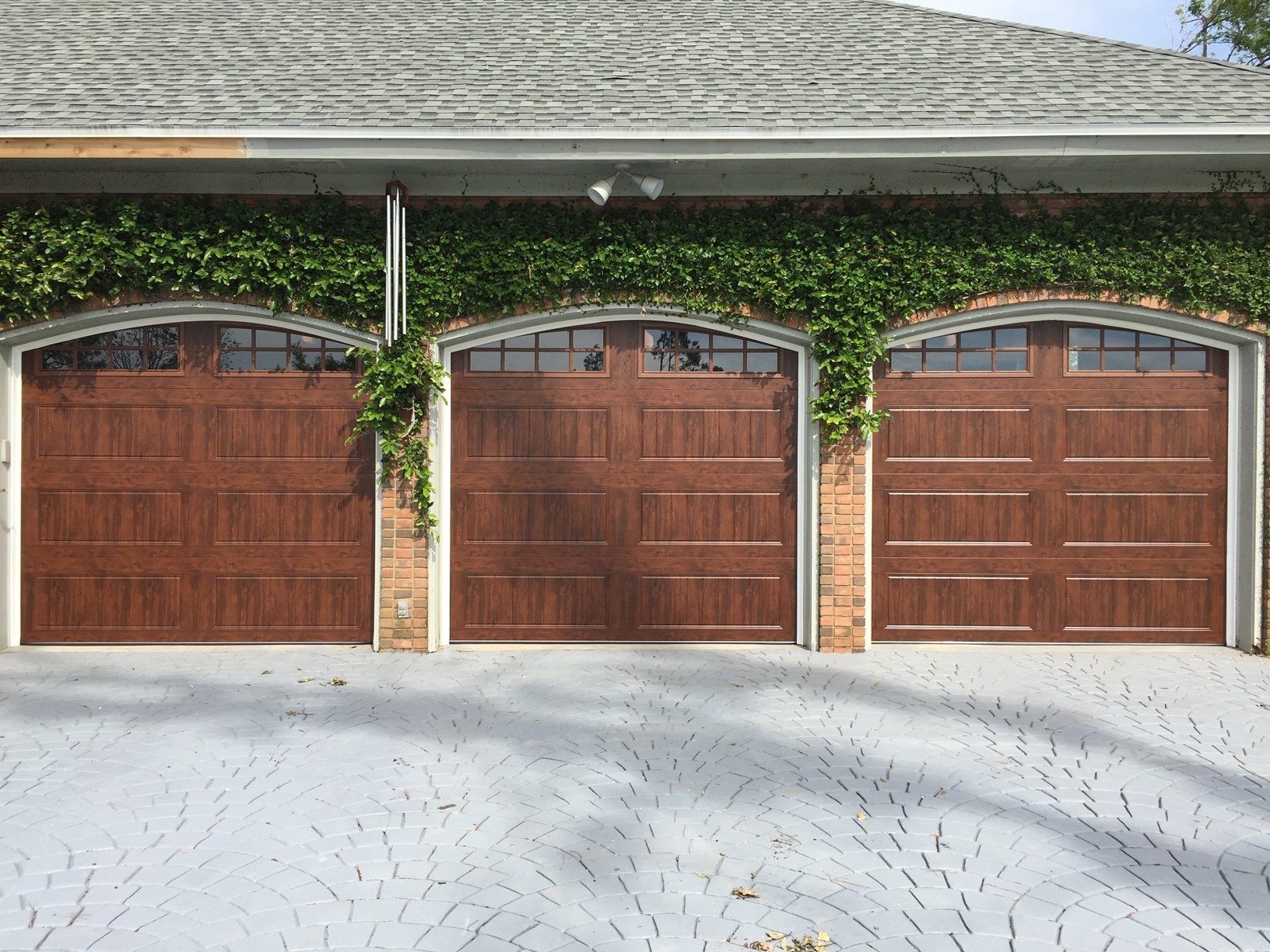 Three brown garage doors with arched windows, brick detailing, and climbing vines on a gray patterned driveway.