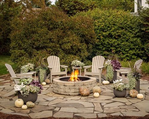 There is a fire pit in the middle of the patio surrounded by chairs and pumpkins.