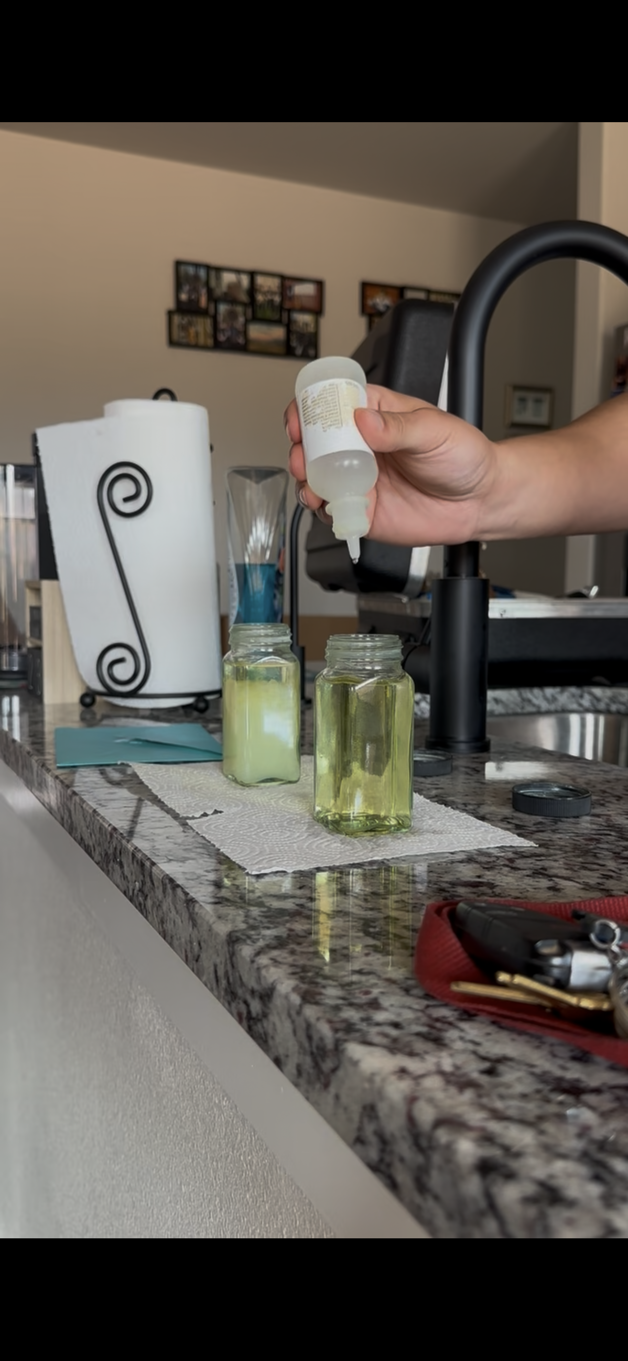 A person is squeezing liquid from a small bottle into a jar on a kitchen counter.