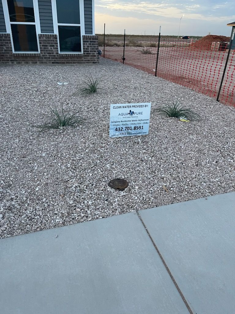 A gravel yard with a sign and a building in the background. A sidewalk is in the foreground.