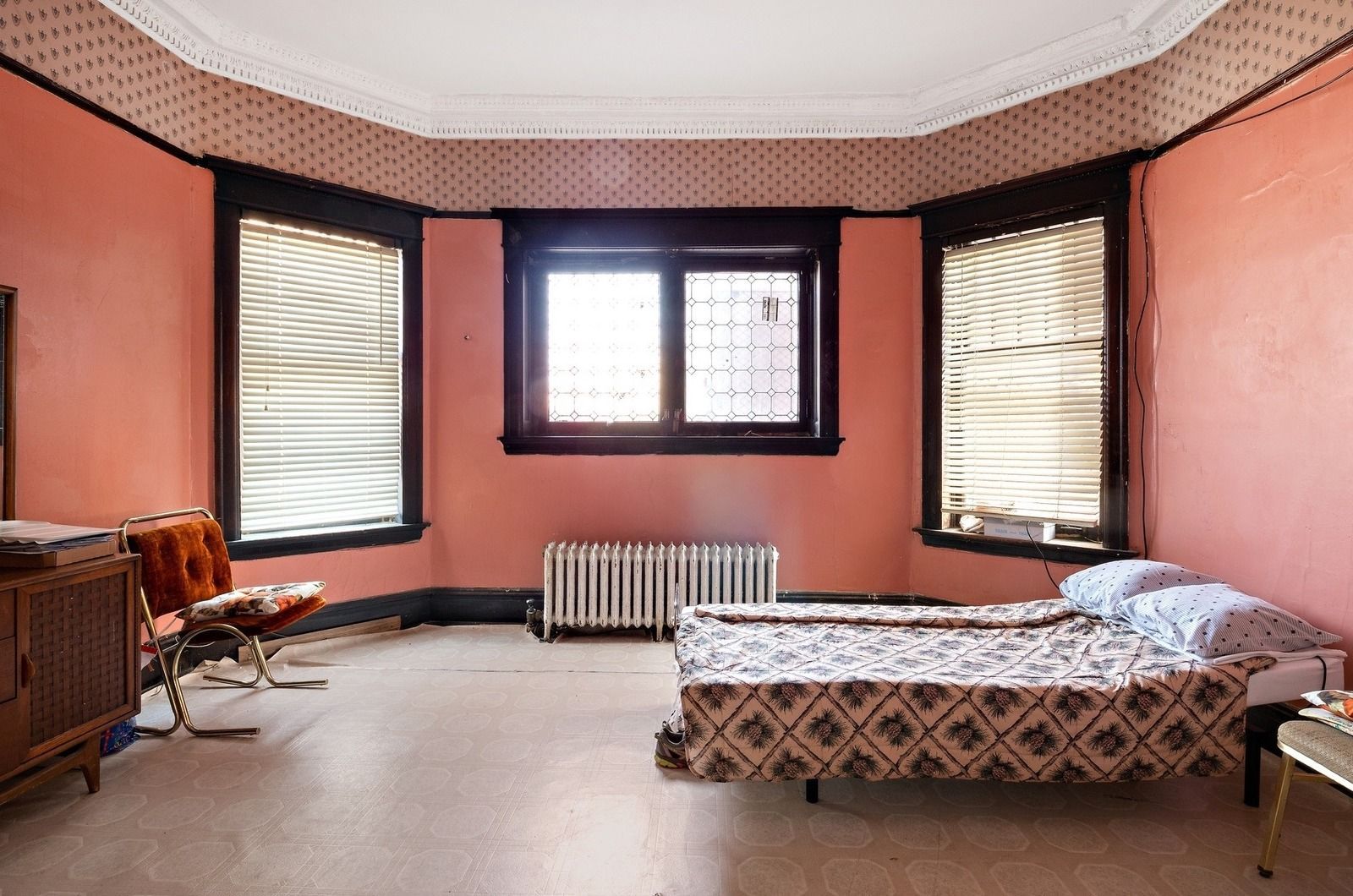 Empty bedroom with pink walls, three windows, a radiator, and a bed by the right wall.