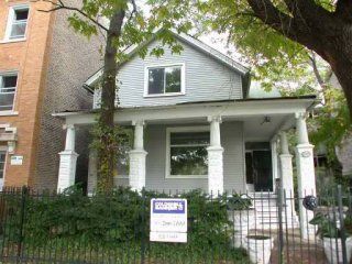 Gray house with white columns and front porch, behind a black fence and sign in front of a tree-lined sidewalk