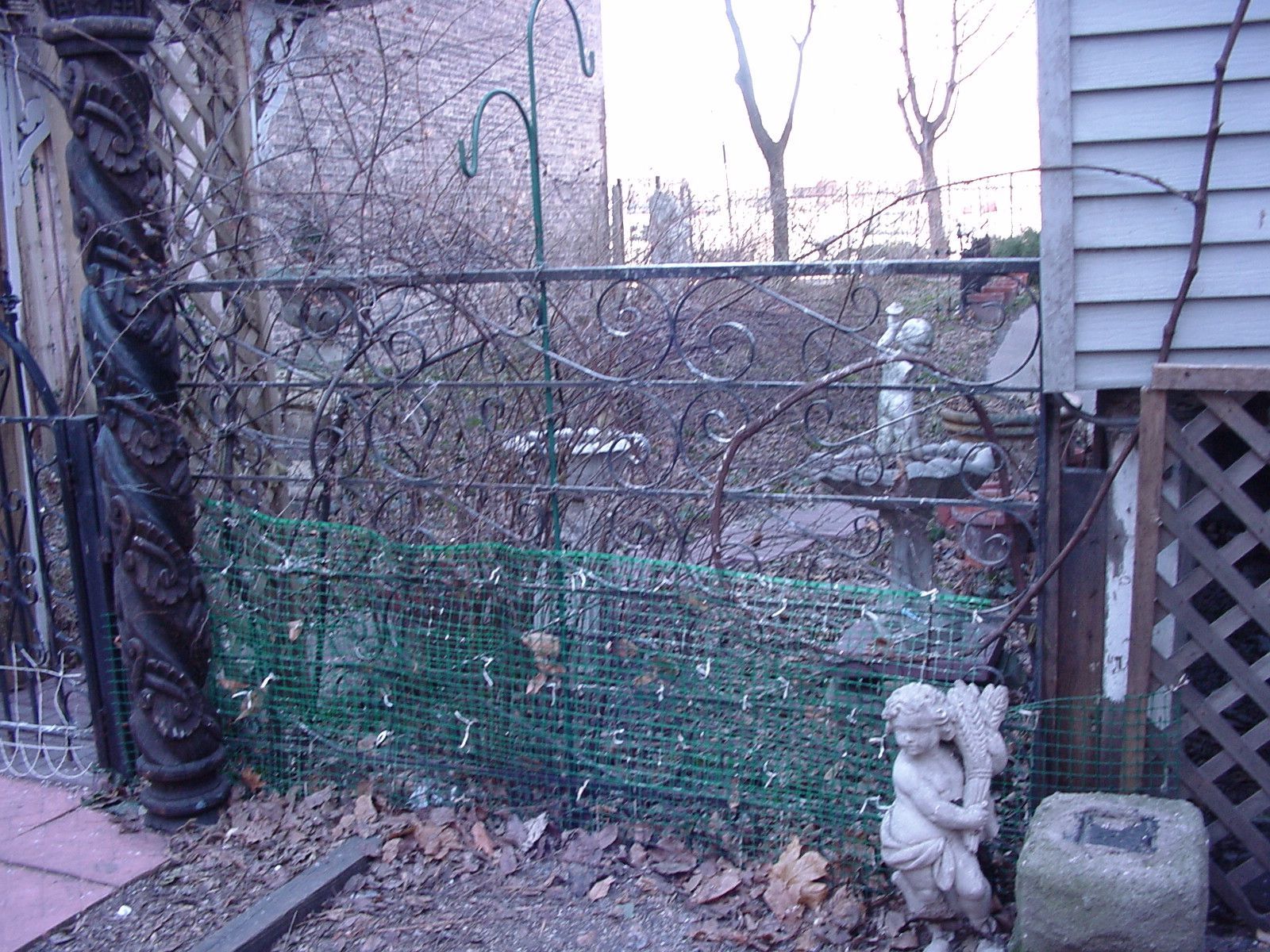Broken green fence beside a house, with bare trees and a small stone dog statue in the yard.