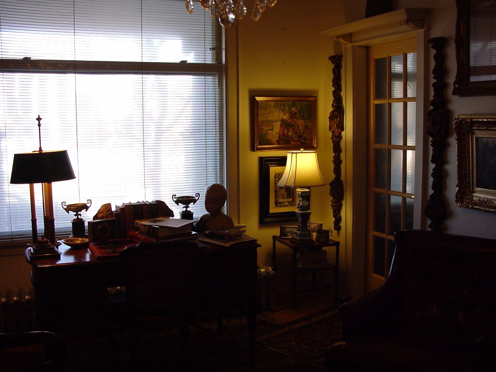 Dimly lit room with desk, lamps, framed art, and sunlight through blinds