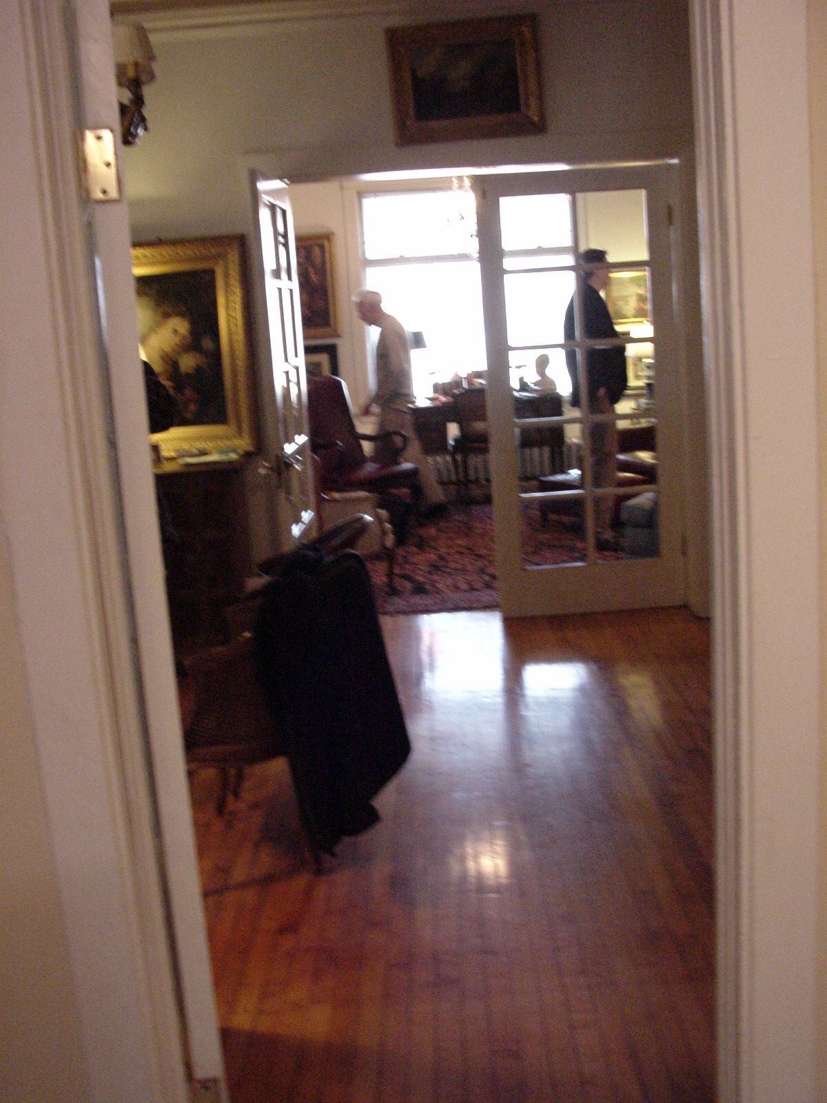Hallway view into a room with hardwood floors, framed art, and two people near a bright window.