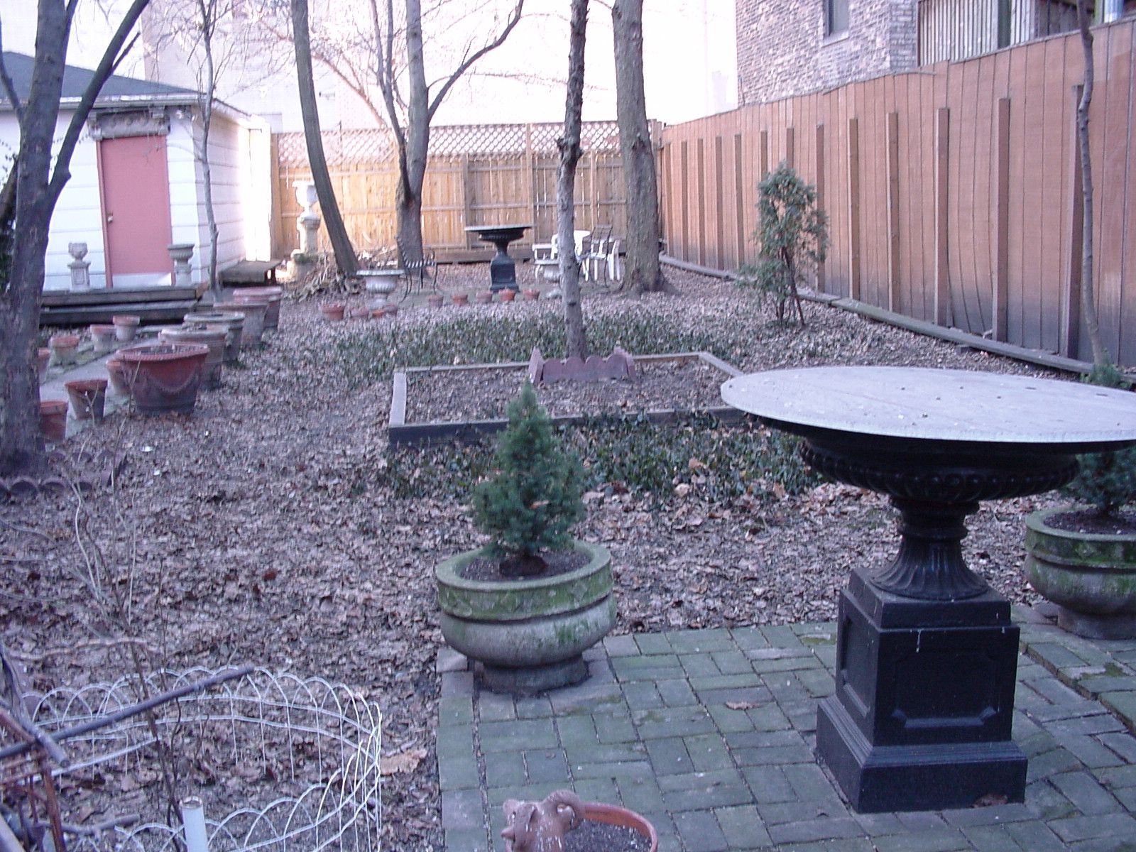 Backyard garden with a birdbath, potted plants, stone path, and wooden fence in winter.
