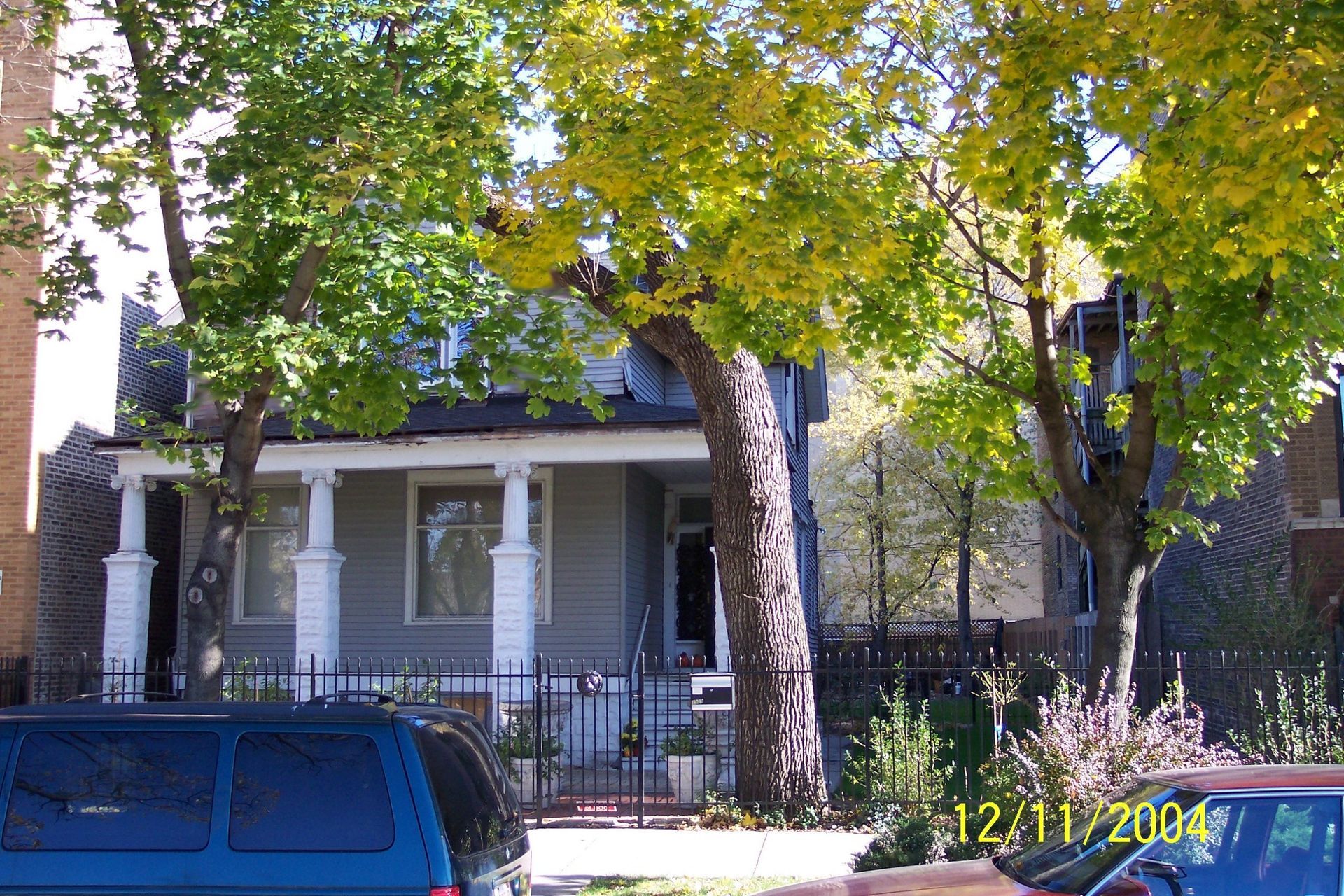 Small gray house with a front porch behind trees and parked cars on a sunny street.