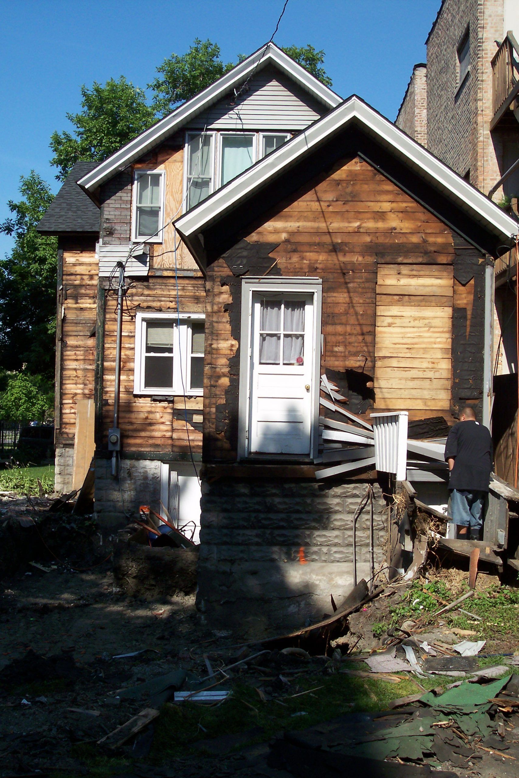 Small wooden house with a white door, boarded window, and debris in the yard under a blue sky