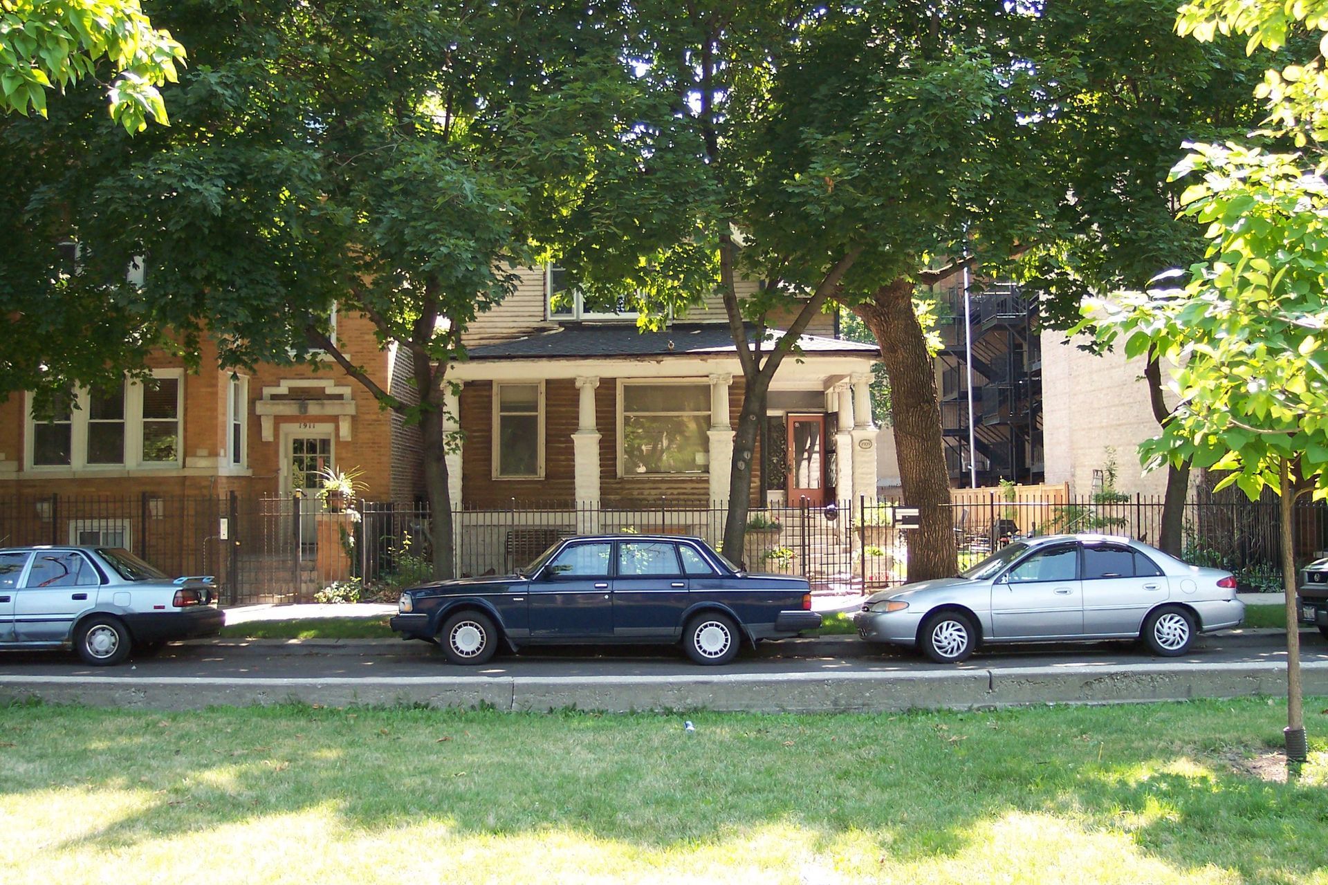 Tree-lined street with parked cars in front of a brick house and porch