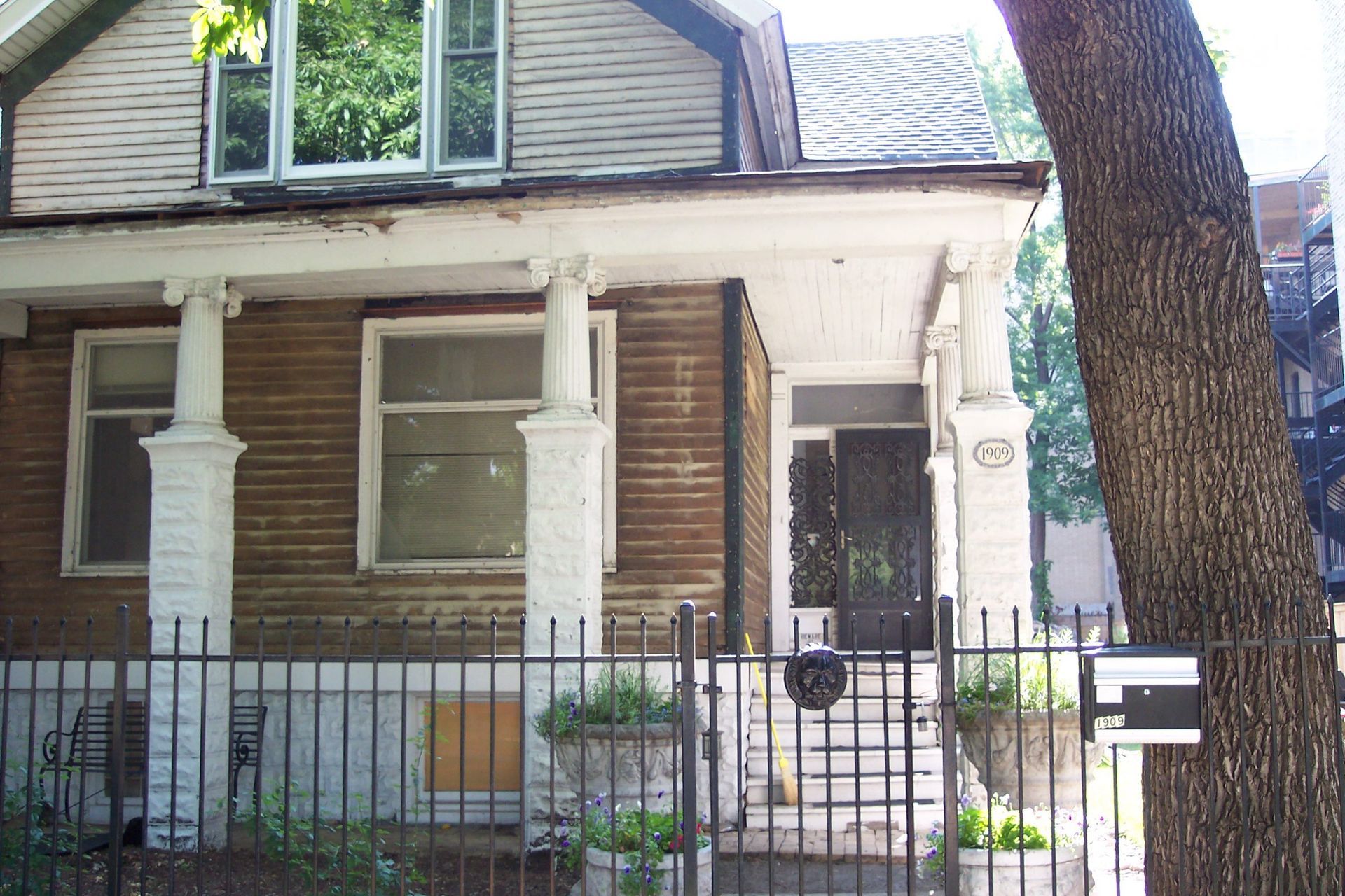 Old brick house with white columns, front porch, iron fence, and a large tree by the entrance