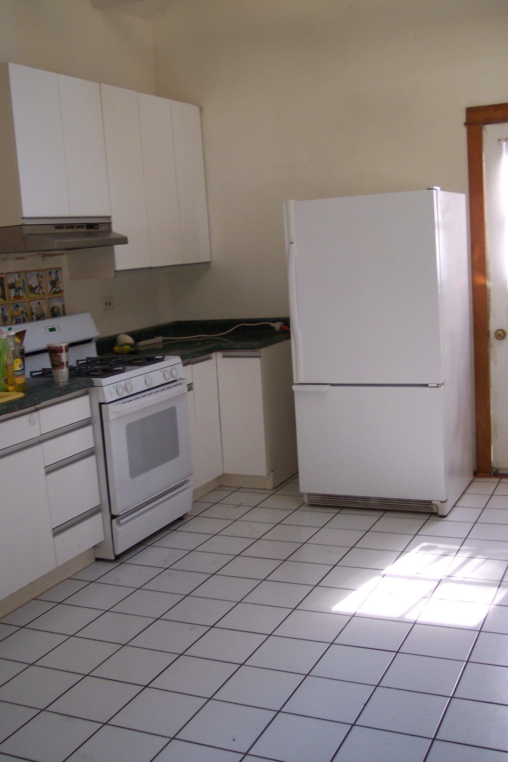 Small kitchen with white cabinets, stove, refrigerator, and tiled floor in sunlight.