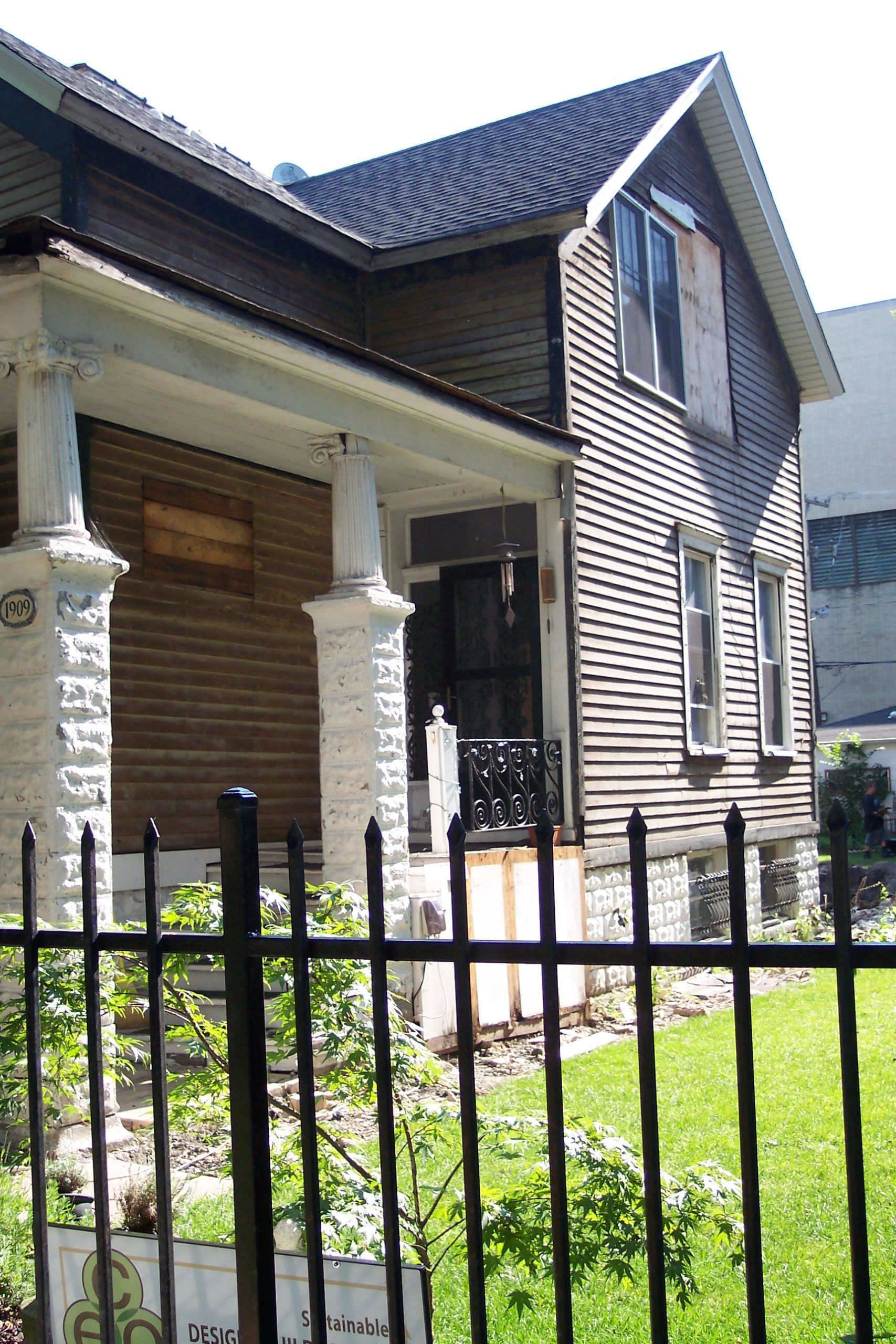 Stone house with dark siding and front porch behind a black fence, in a sunny yard