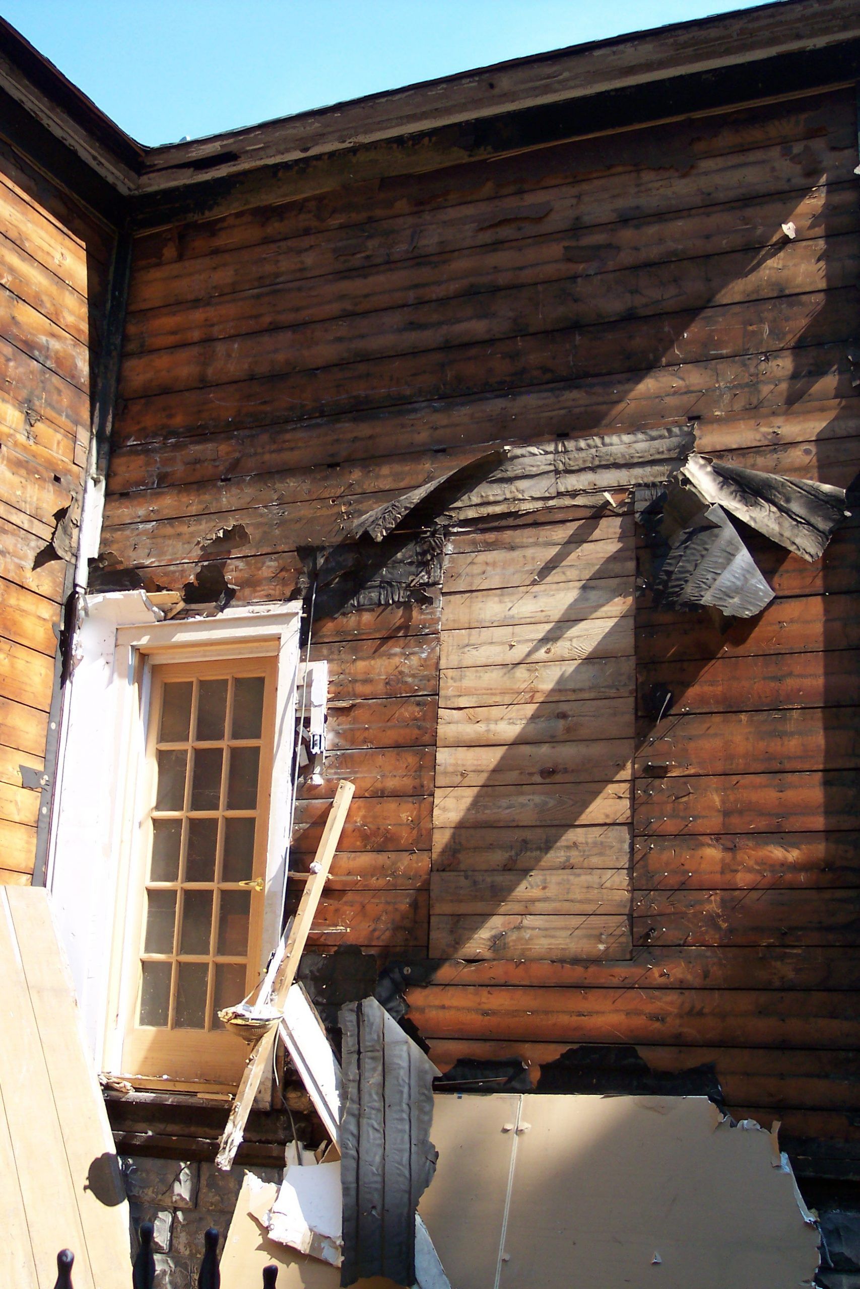 Damaged wooden house wall with torn siding and a broken window in bright sunlight