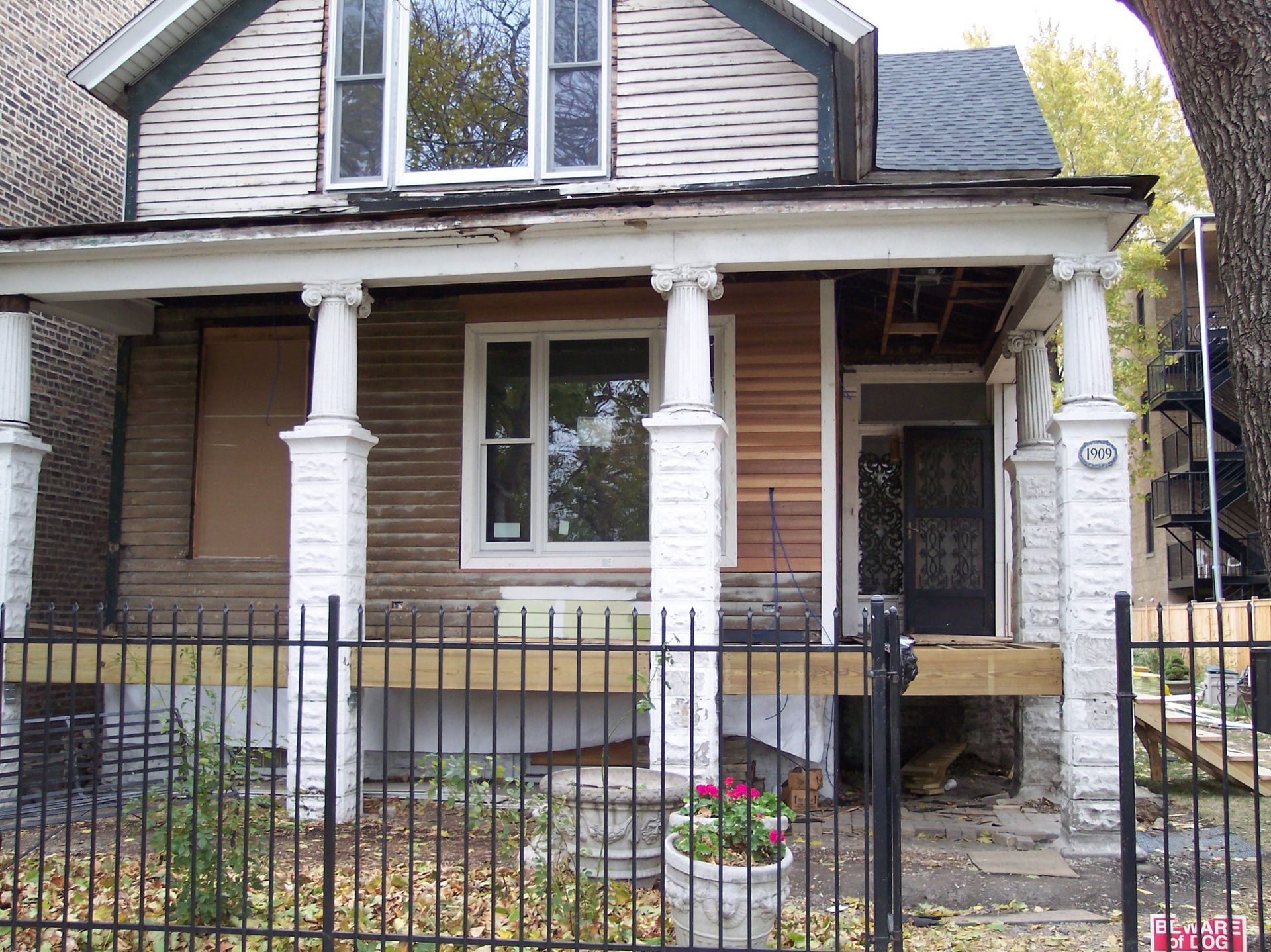 Front porch of a weathered house with white columns, black fence, and a small flower planter