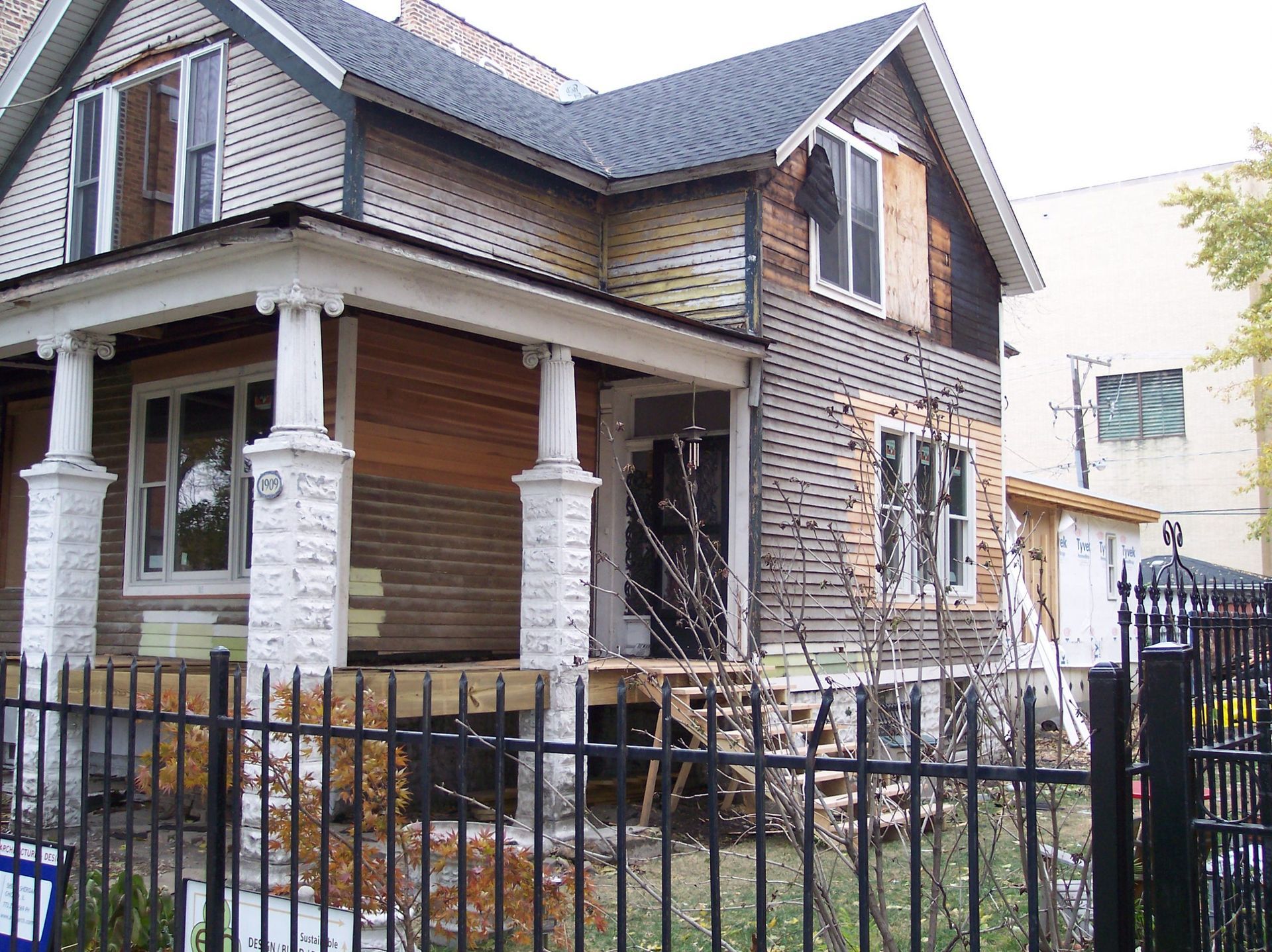 Damaged two-story house with a boarded porch and broken windows behind a black metal fence