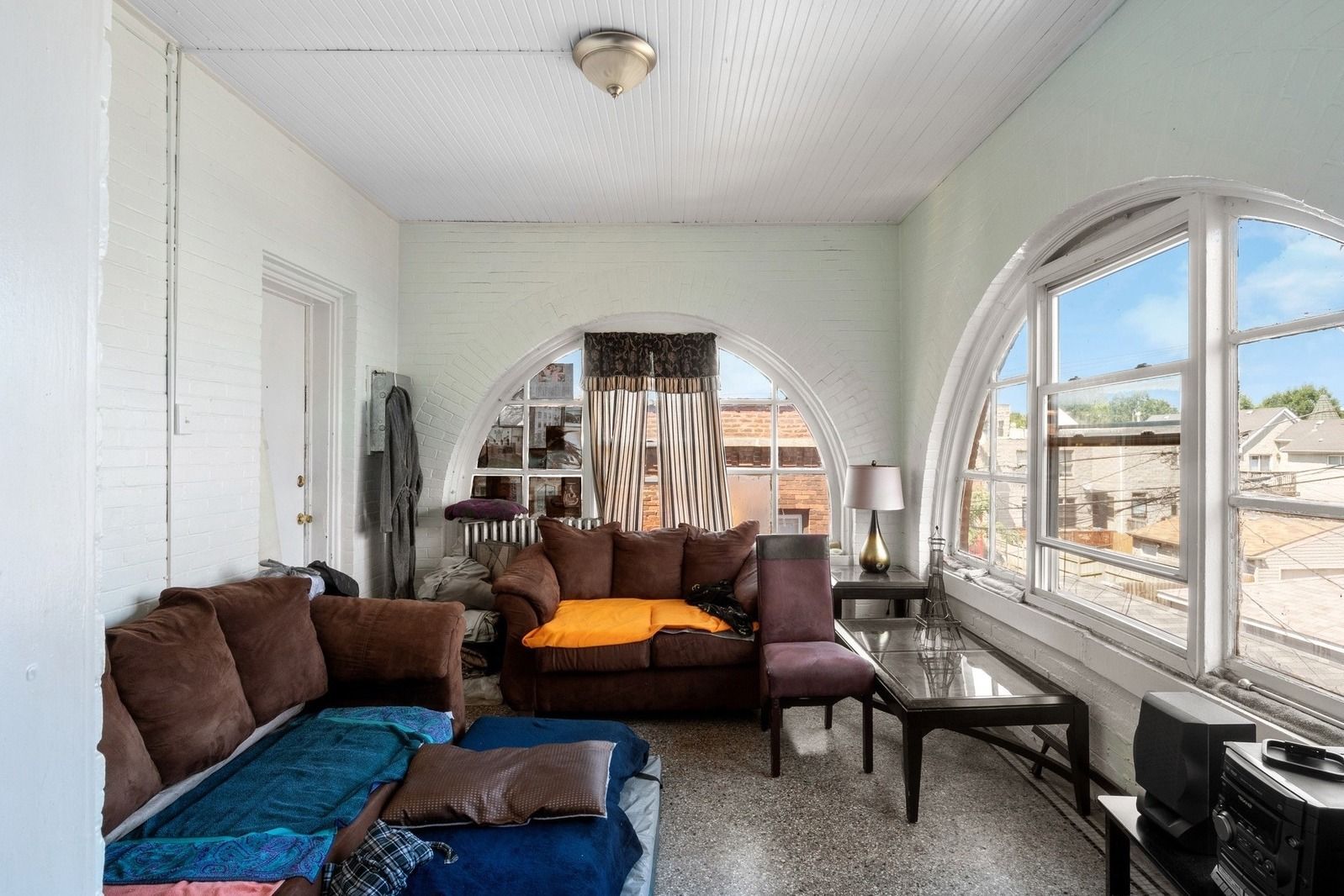 Bright living room with brown sofa, orange chair, and large arched window overlooking rooftops.