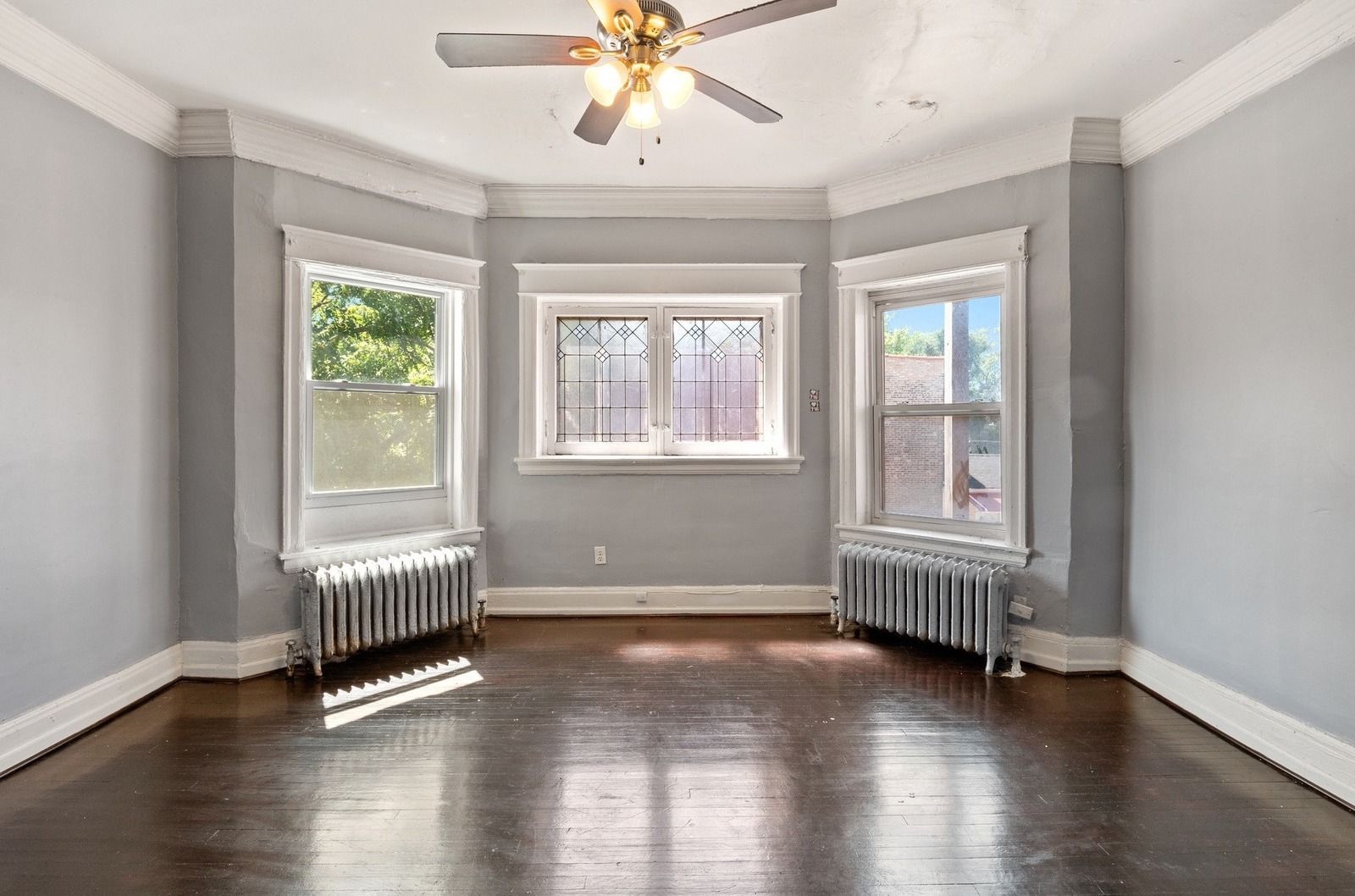 Empty gray living room with three windows, hardwood floors, and a ceiling fan