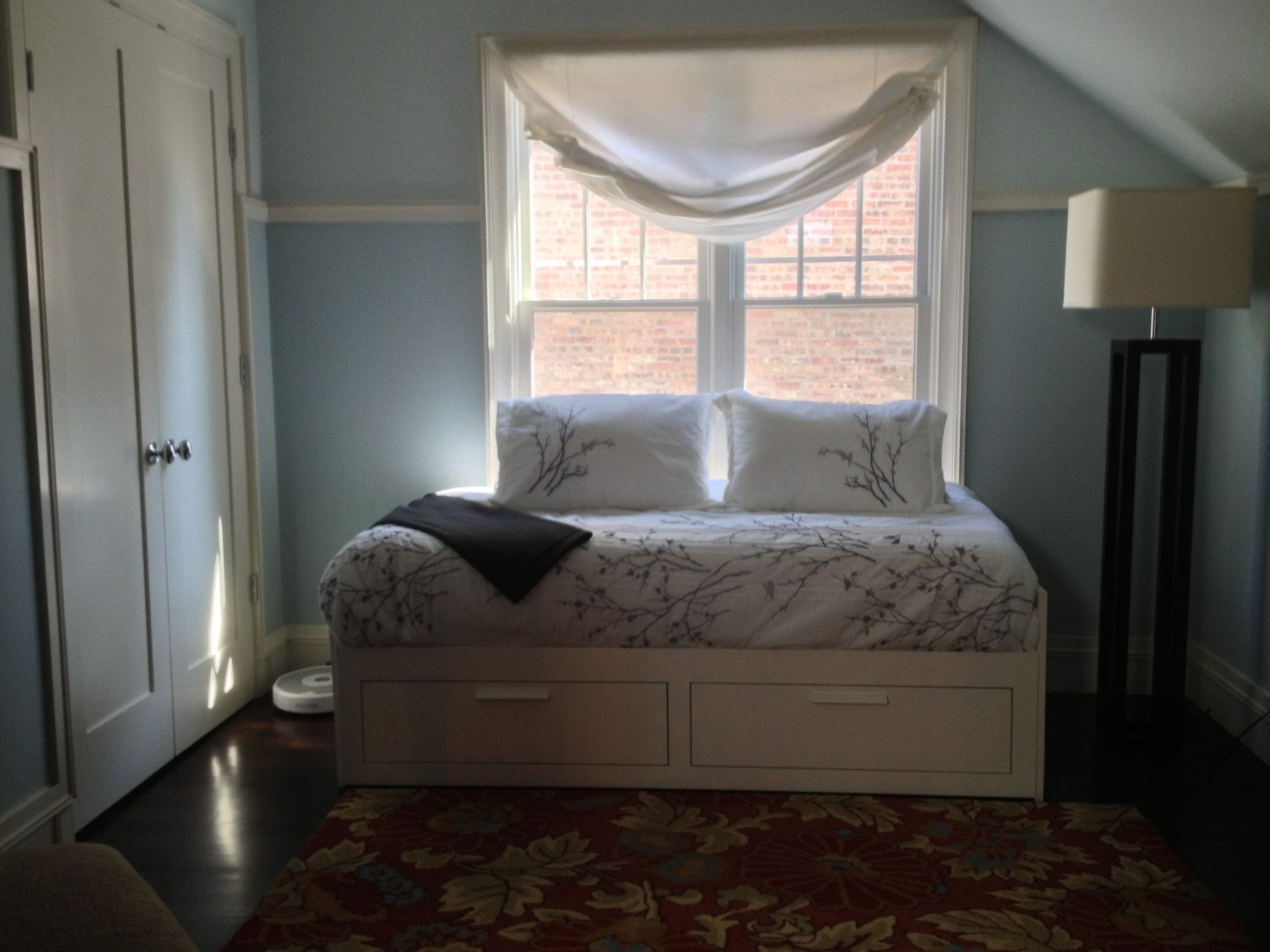 Small bedroom with a bed by a window, white curtains, a lamp, and a floral rug.