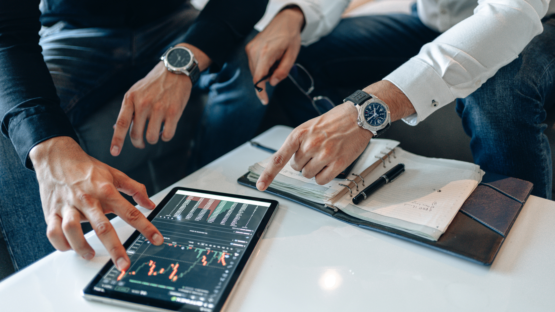 Three people in business attire review charts on a tablet and notebook at a table.