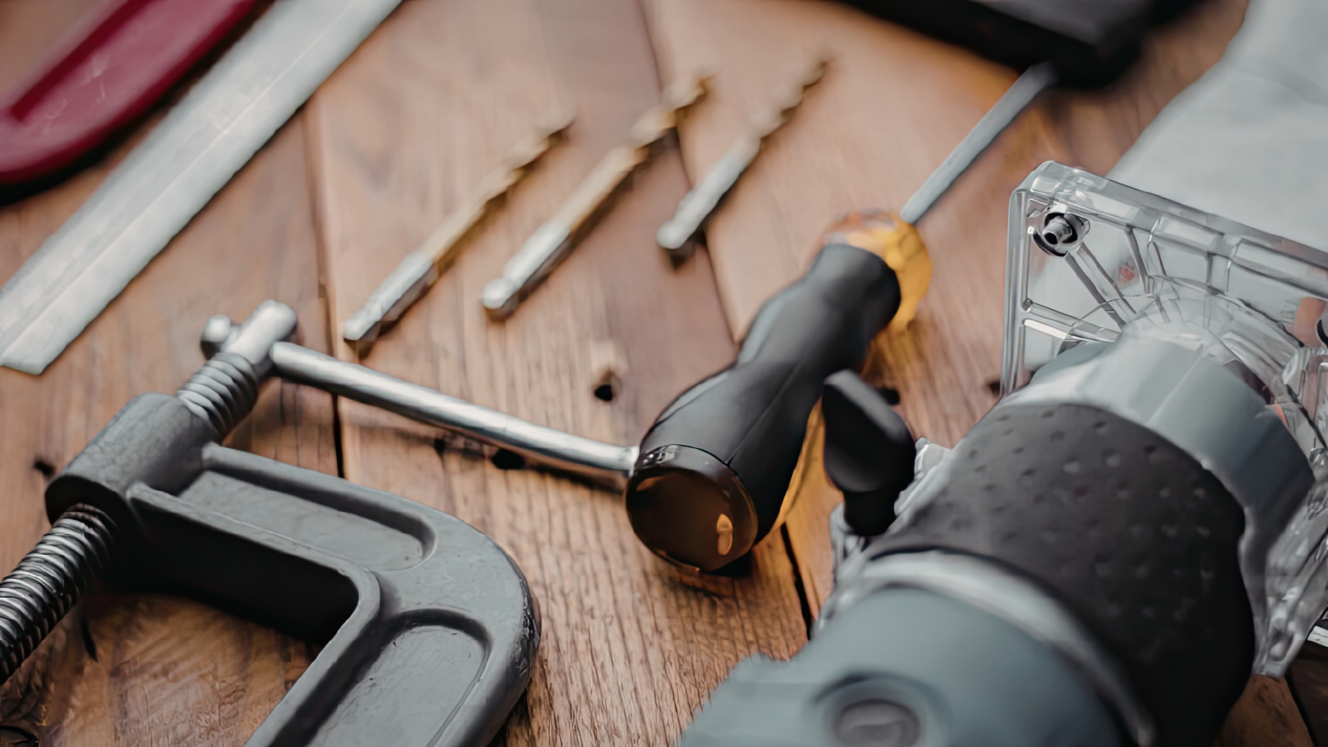 Hand tools and drill bits on a wooden workbench, with a clamp and cordless drill in view