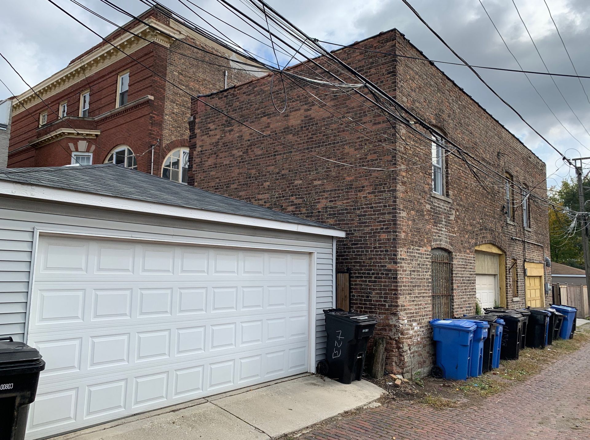 Brick alley beside a white garage, with blue trash bins and overhead utility wires