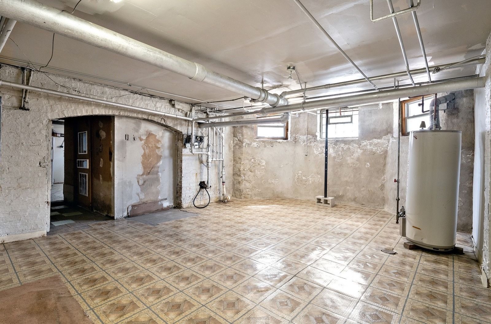 Empty unfinished basement with white walls, tiled floor, exposed pipes, and a furnace unit.