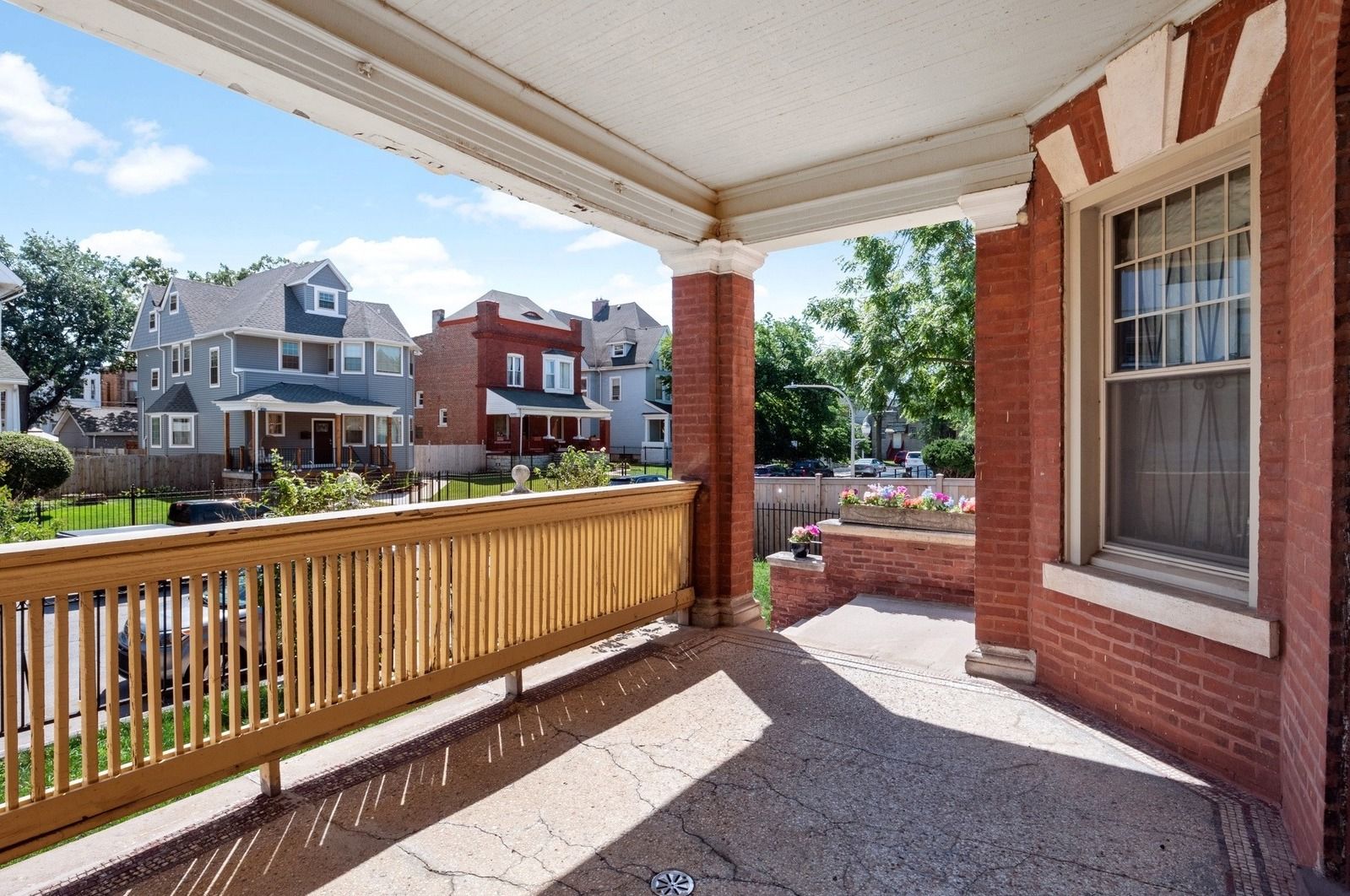 Covered front porch with brick columns, wooden railing, and neighborhood houses beyond.