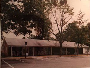 Long, low, brick building with a covered porch and chairs, in a parking lot with trees.
