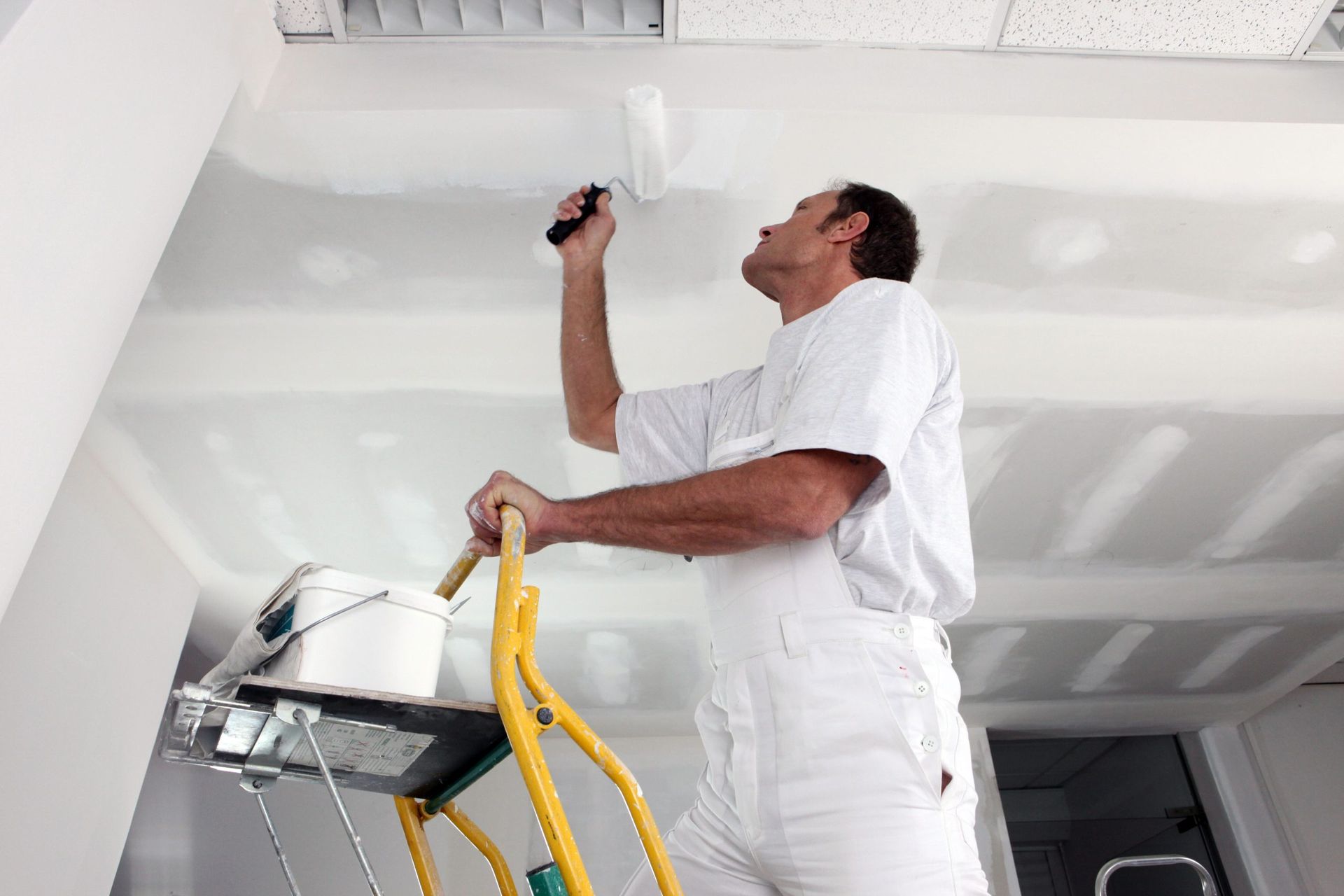 Painter on a ladder painting a white ceiling with a roller. Interior setting.