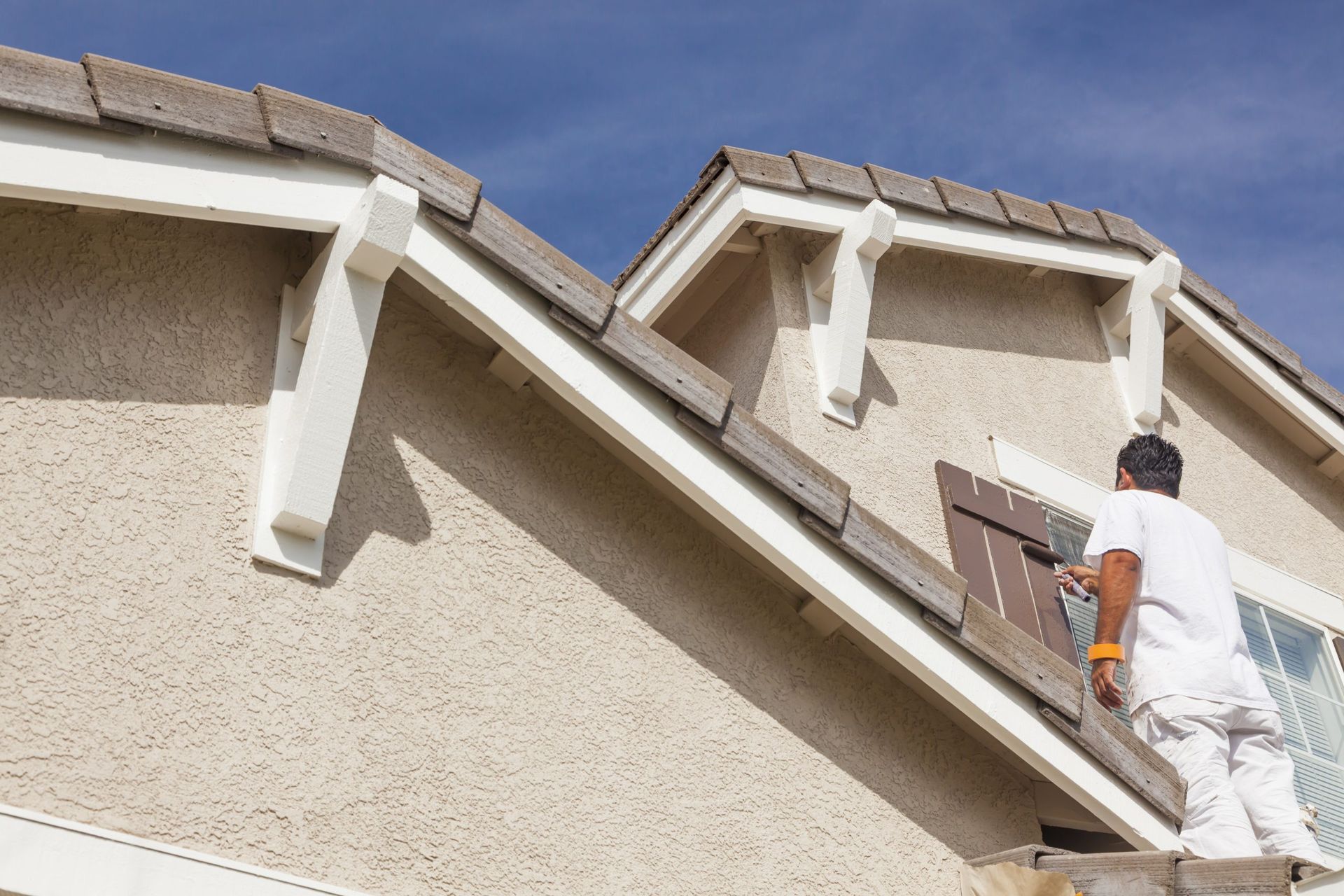 Person on a ladder painting a house's trim and eaves; clear blue sky in the background.
