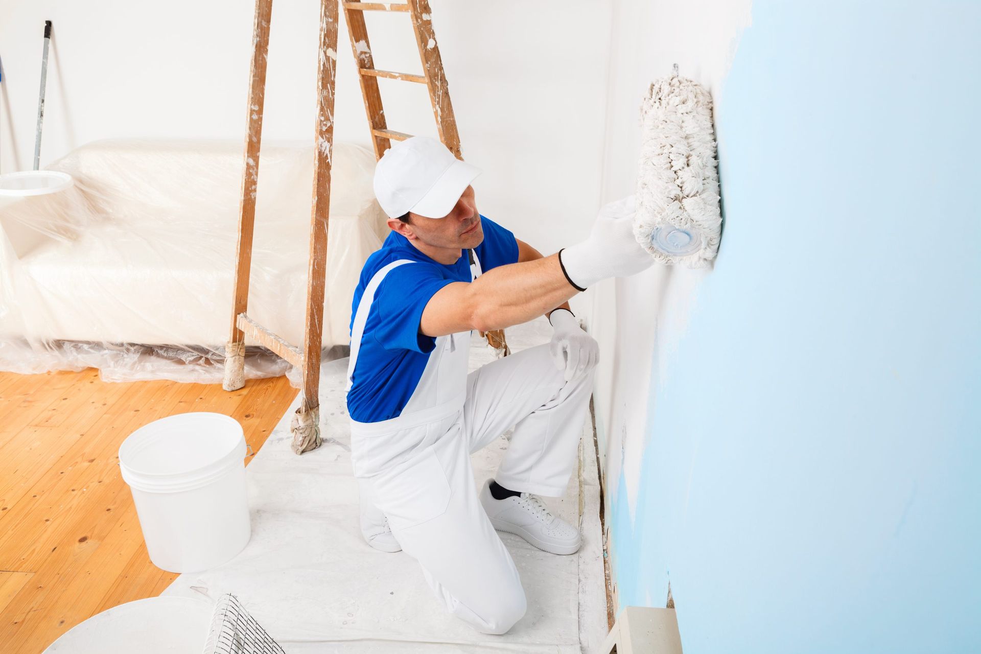 Painter kneeling, rolling light blue paint onto a white wall. Wooden ladder and paint bucket in the background.