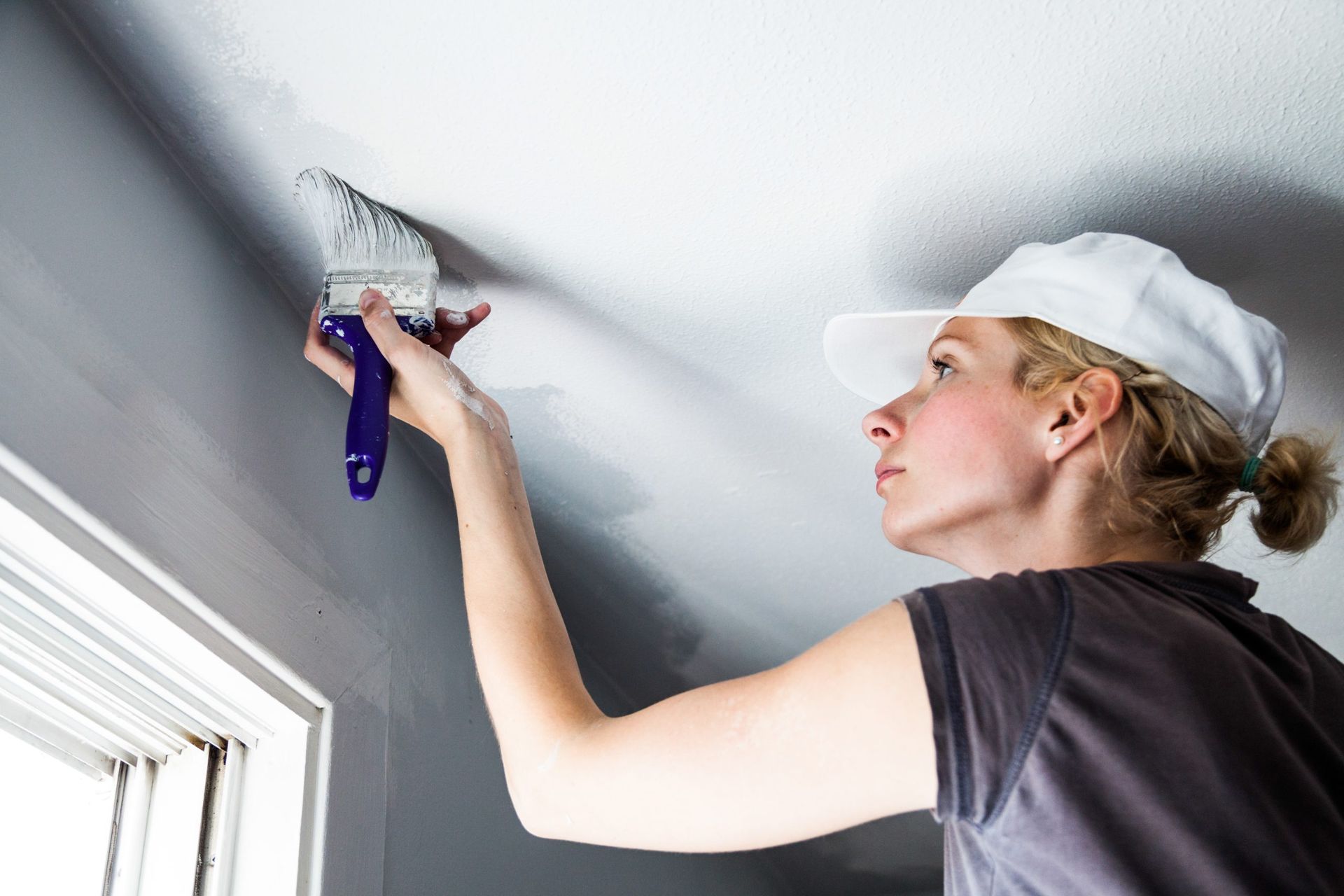 Woman paints a ceiling white with a brush, standing near a window.