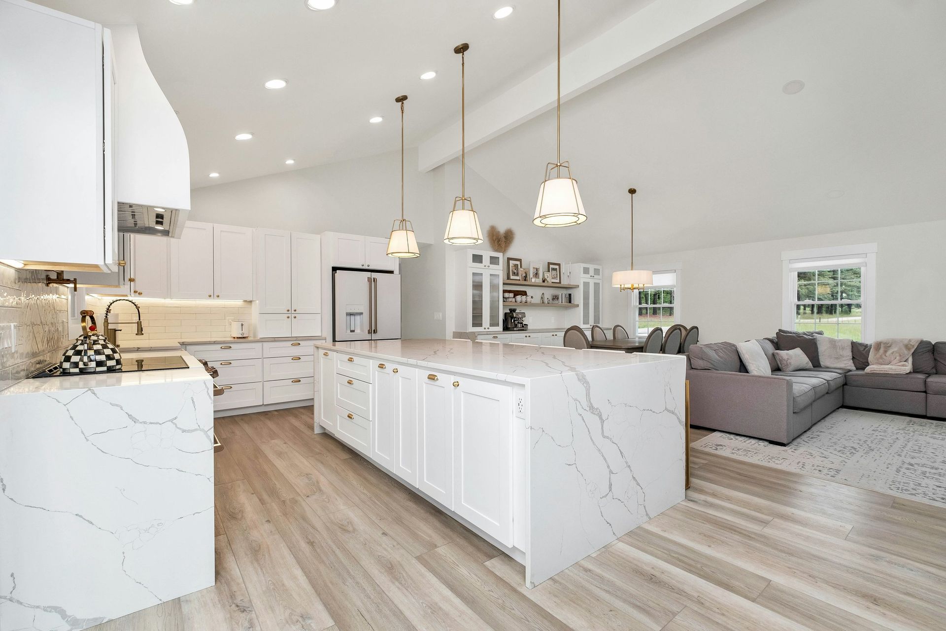 White kitchen with large island and light wood floors, leading to a living area with a gray sectional.