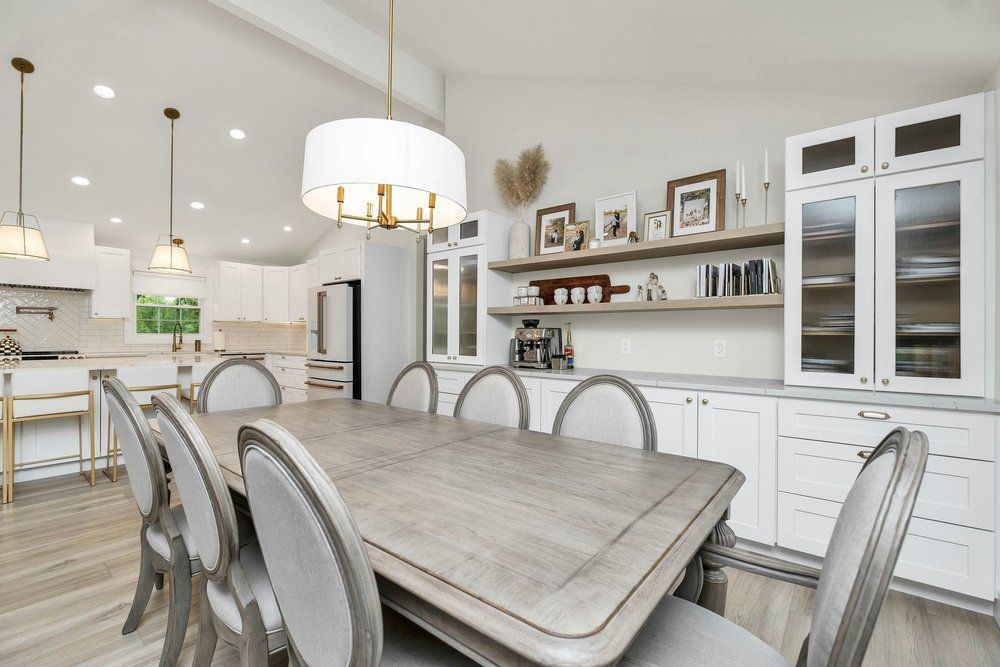 Elegant white dining room with a long table and eight upholstered chairs, kitchen in background.