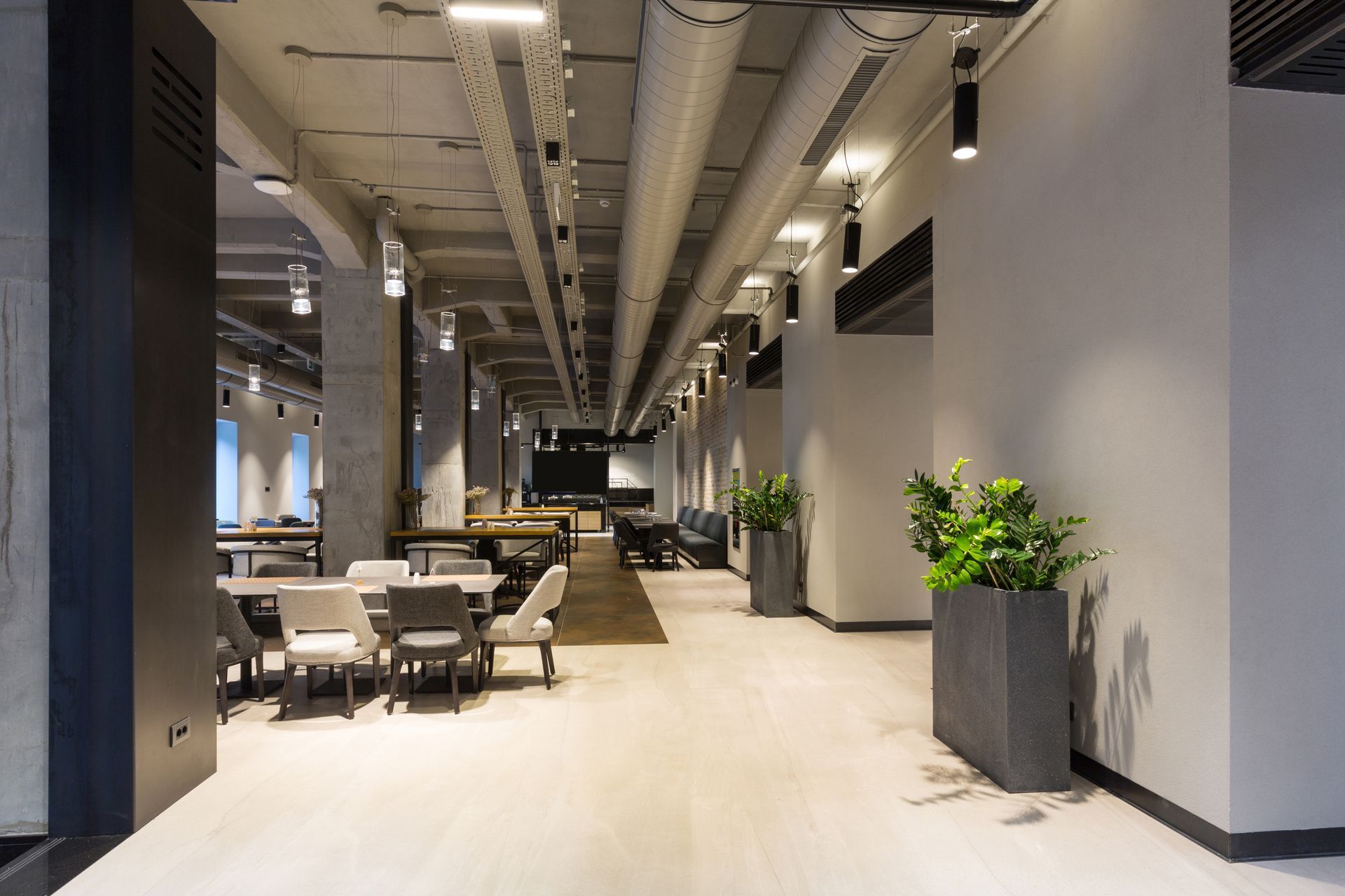 Long, modern hallway with tables, chairs, and potted plants. Exposed pipes and concrete ceiling.