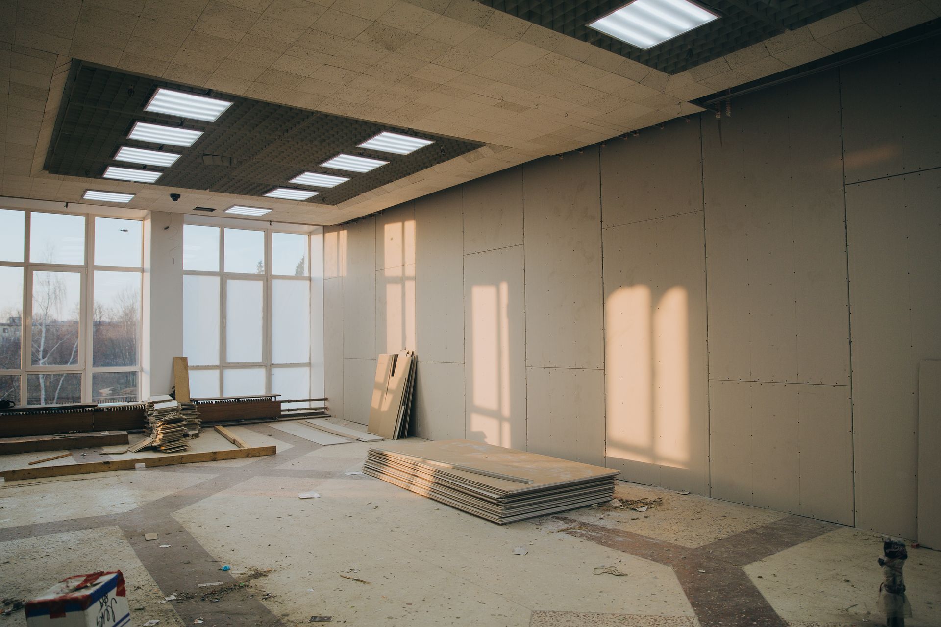Interior of a room under construction with unfinished walls, windows, and debris on the floor. Sunlight.