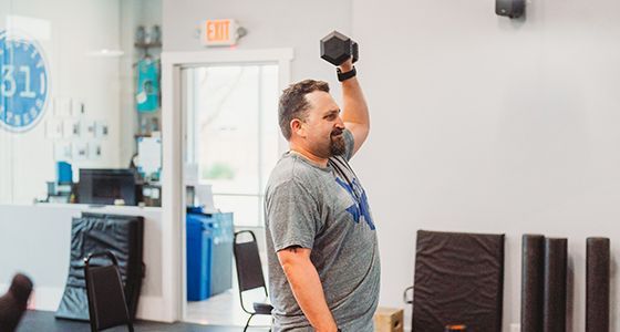 Man lifts a dumbbell over his head in a gym. He is smiling and wearing a gray shirt.